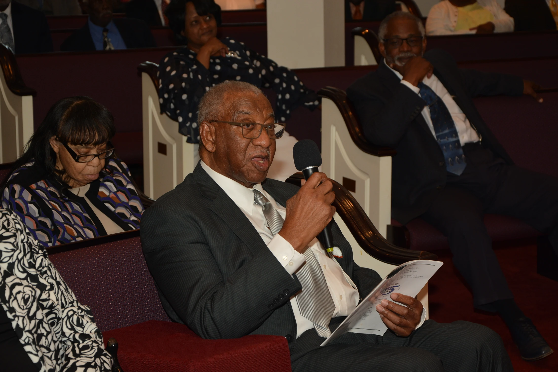 An older Black man in a suit and glasses speaks into a microphone while seated in a pew, holding a program. Other people are seated in the background.