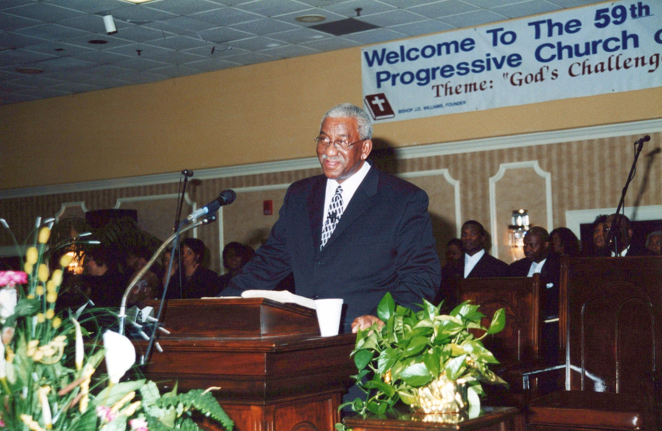 An older Black man in a suit and glasses smiles while speaking at a wooden lectern. A banner for the 59th Progressive Church conference hangs behind him.