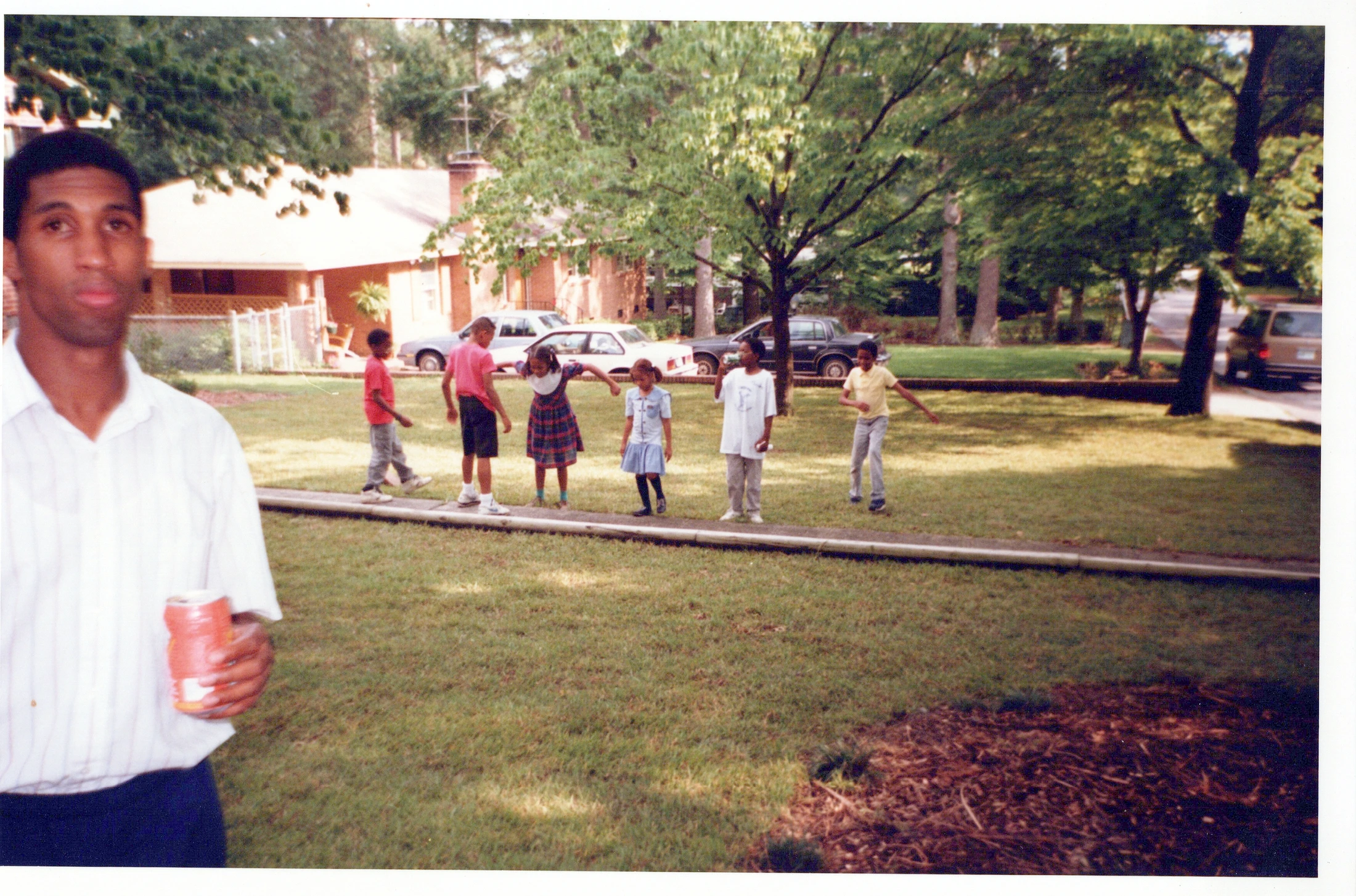 A vintage photo of a Black man in the foreground holding a can, with several Black children playing on a grassy lawn behind him in a suburban neighborhood.