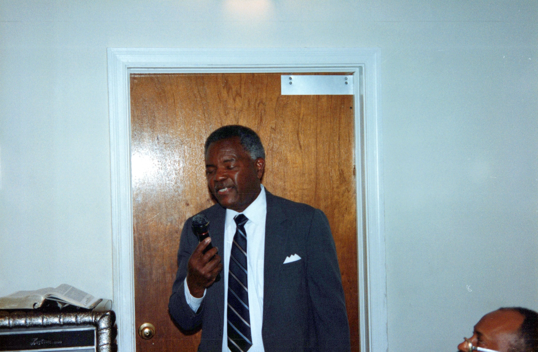 A film photograph of an older Black man in a grey suit and striped tie. He stands indoors in front of a wooden door, speaking into a microphone.