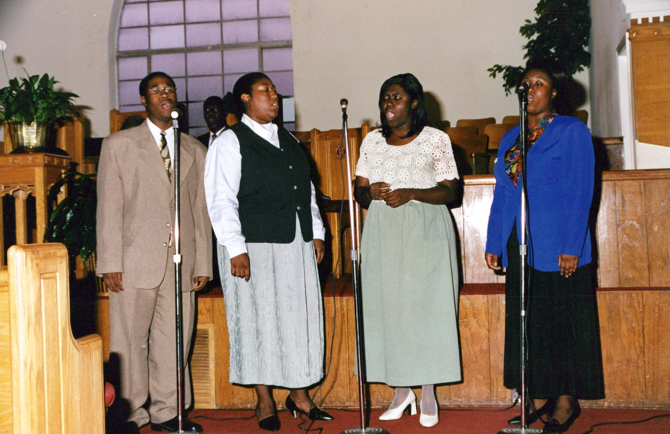A man and three women stand in a line at the front of a church, each singing into a microphone during a performance.