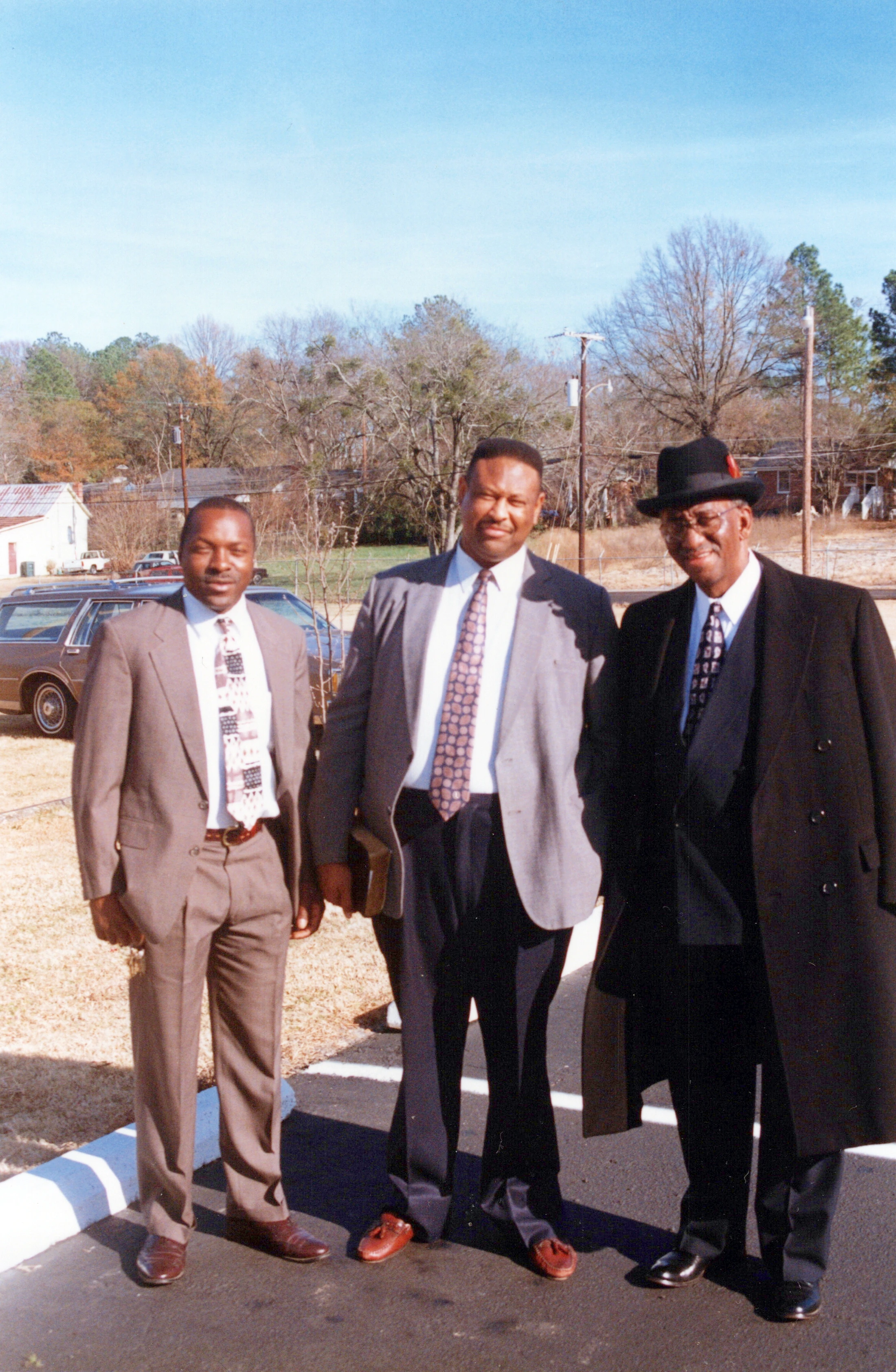 Three Black men in suits and ties stand outside in a parking lot on a sunny day. The man on the right wears a fedora and an overcoat.