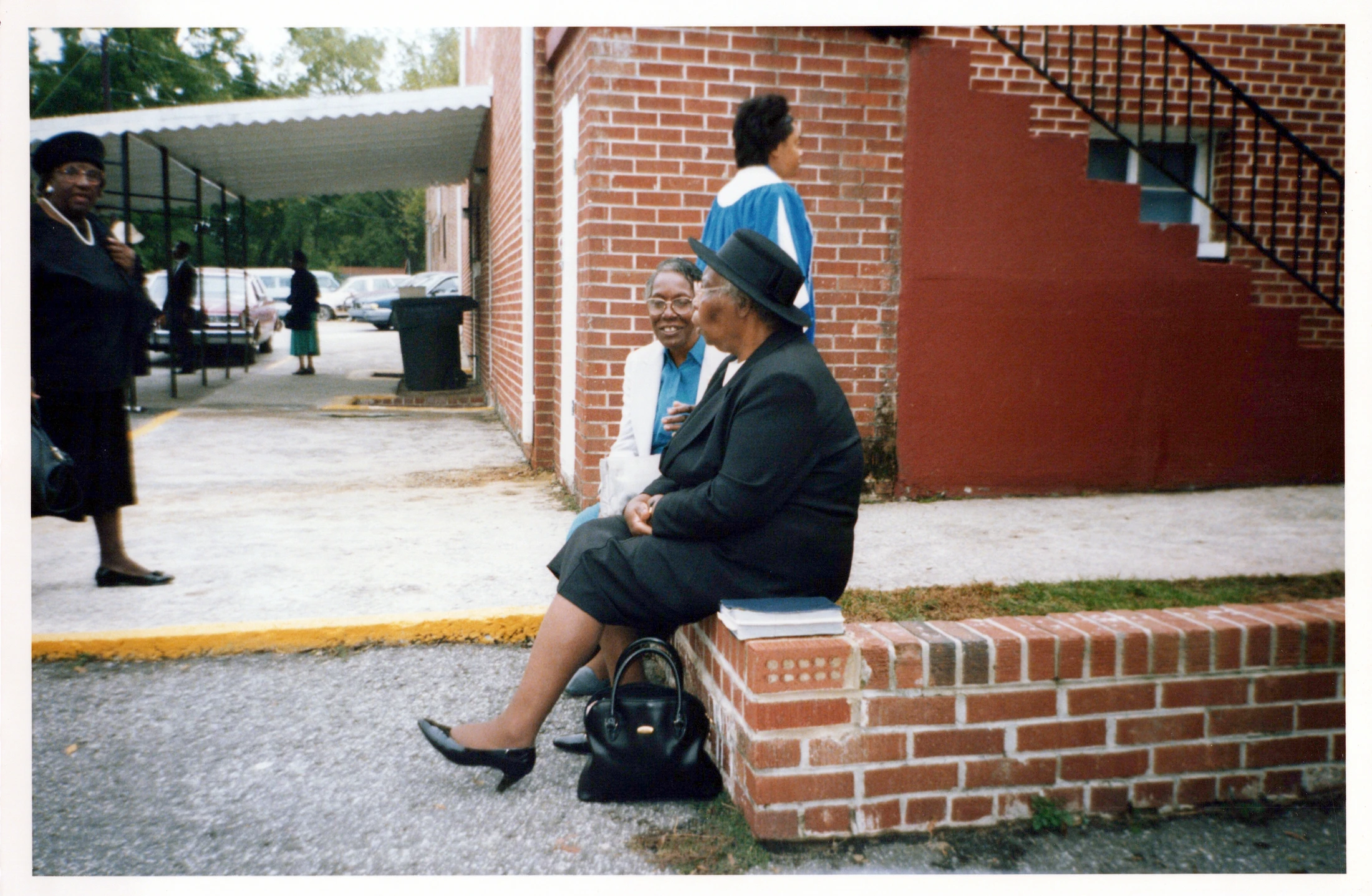 Several Black women in Sunday attire gather outside a red brick building. Two women sit on a low brick wall and chat, while others stand nearby.