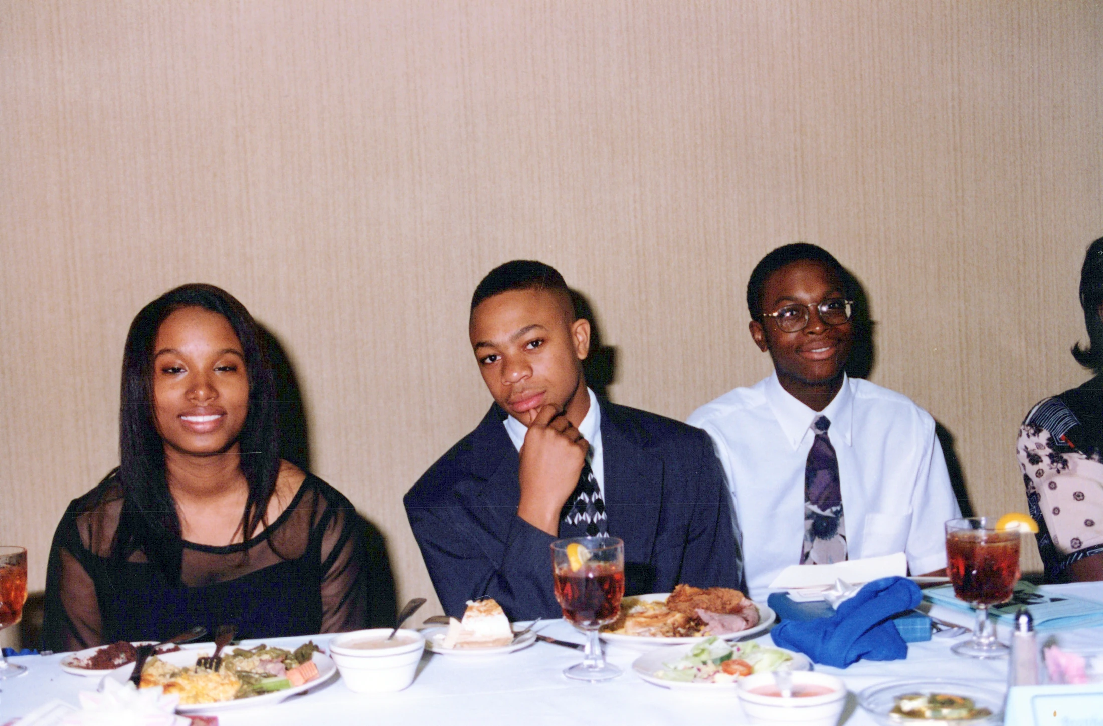 Three formally dressed African American teenagers pose for a photo while seated at a dining table during a banquet. The table is filled with food and drinks.