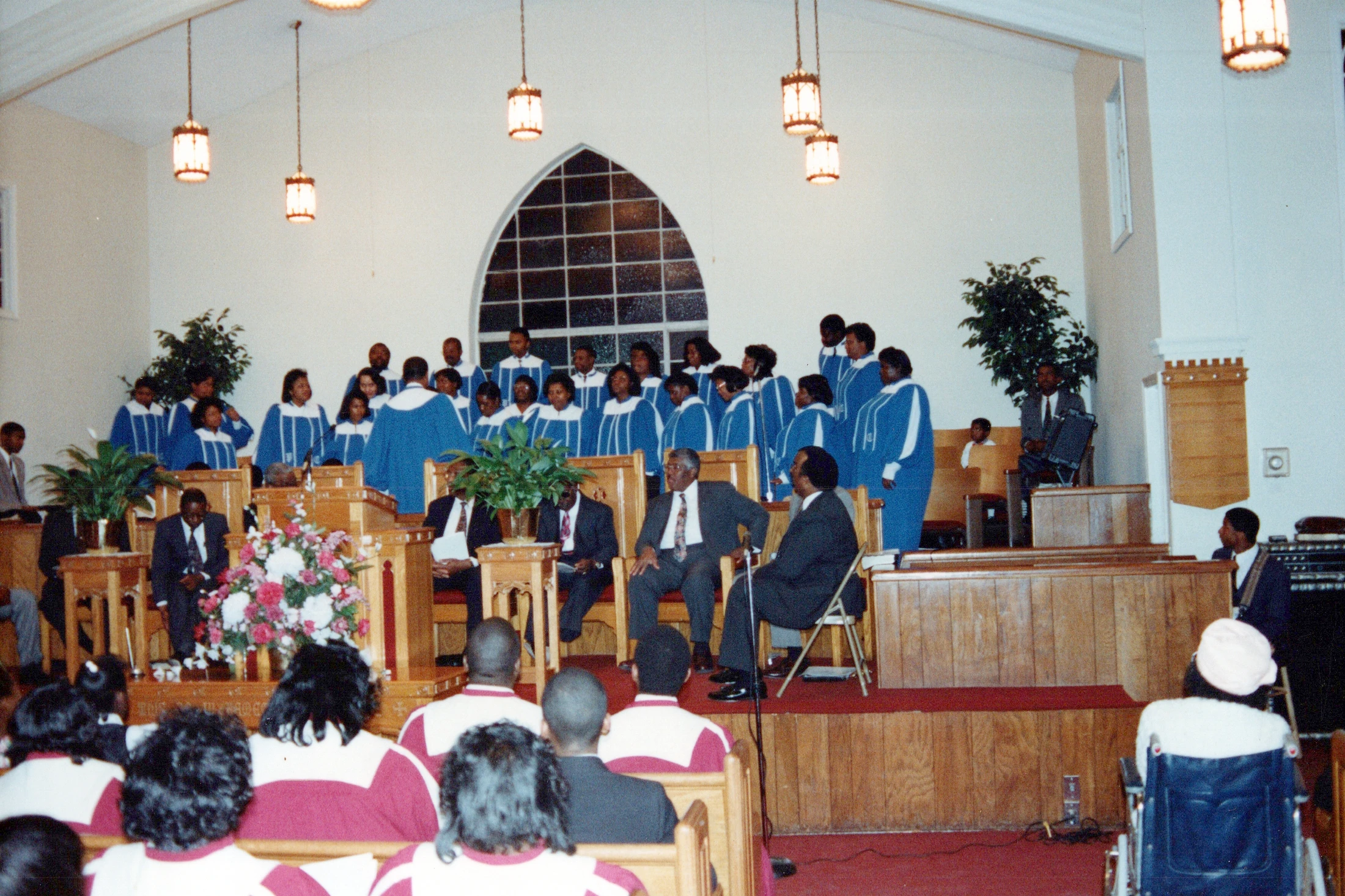 A wide shot of a church sanctuary during a service. A choir in blue and white robes stands on a platform behind several men in suits, facing the congregation.
