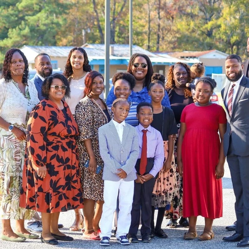 A large, multi-generational Black family poses for a group photo in a sunny parking lot. The men, women, and children are smiling and dressed in formal attire.