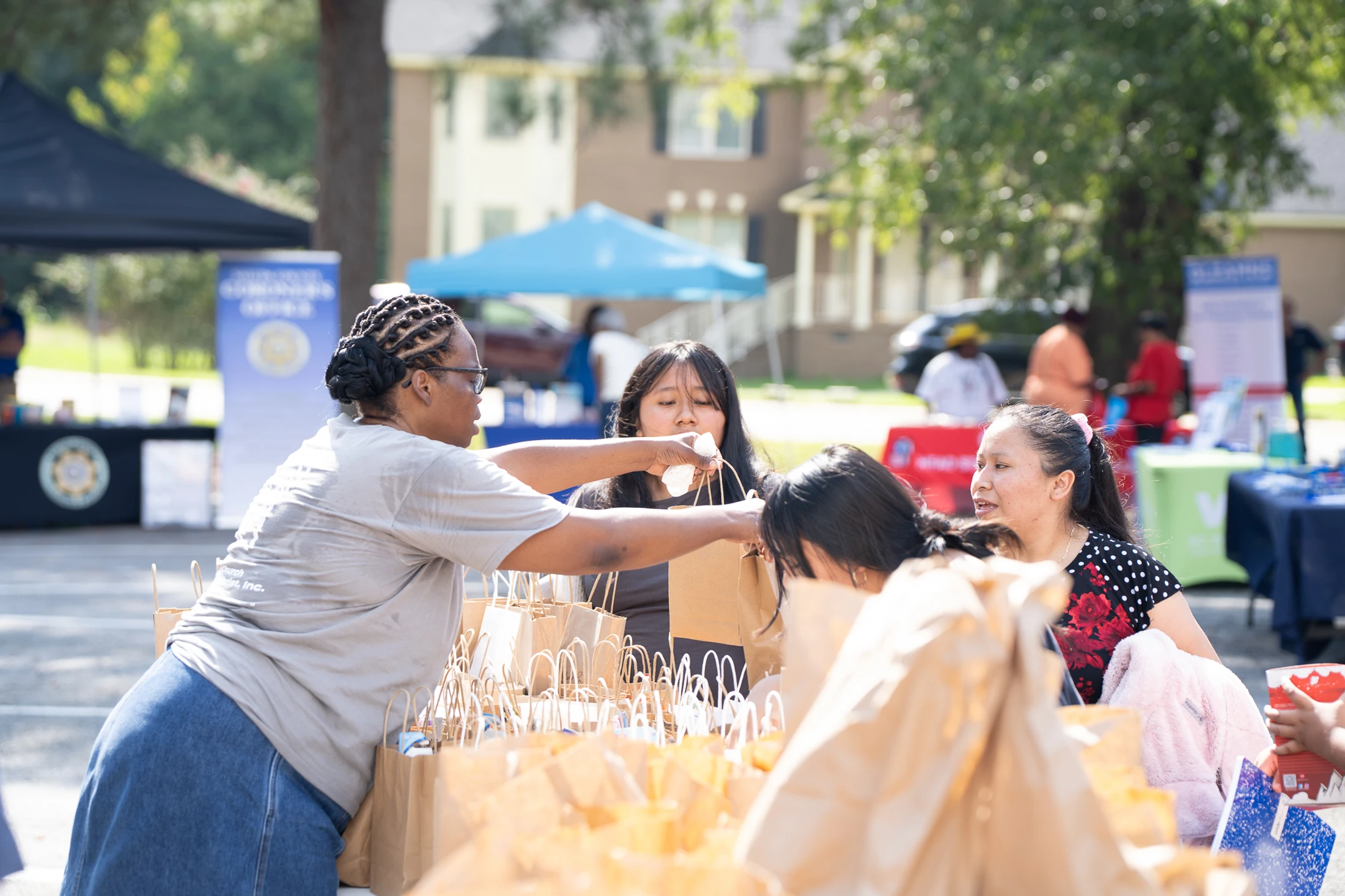 A woman with braided hair hands a paper gift bag to another woman at an outdoor community fair. A table is filled with many similar bags for a giveaway.
