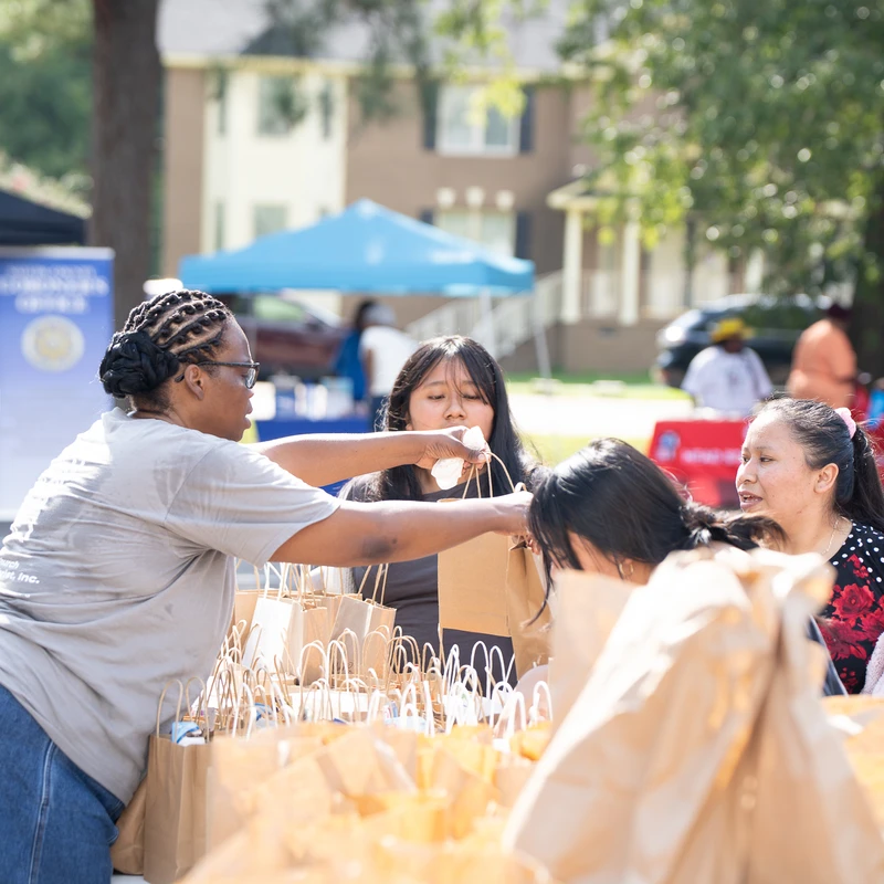 A woman with braided hair hands a paper gift bag to another woman at an outdoor community fair. A table is filled with many similar bags for a giveaway.