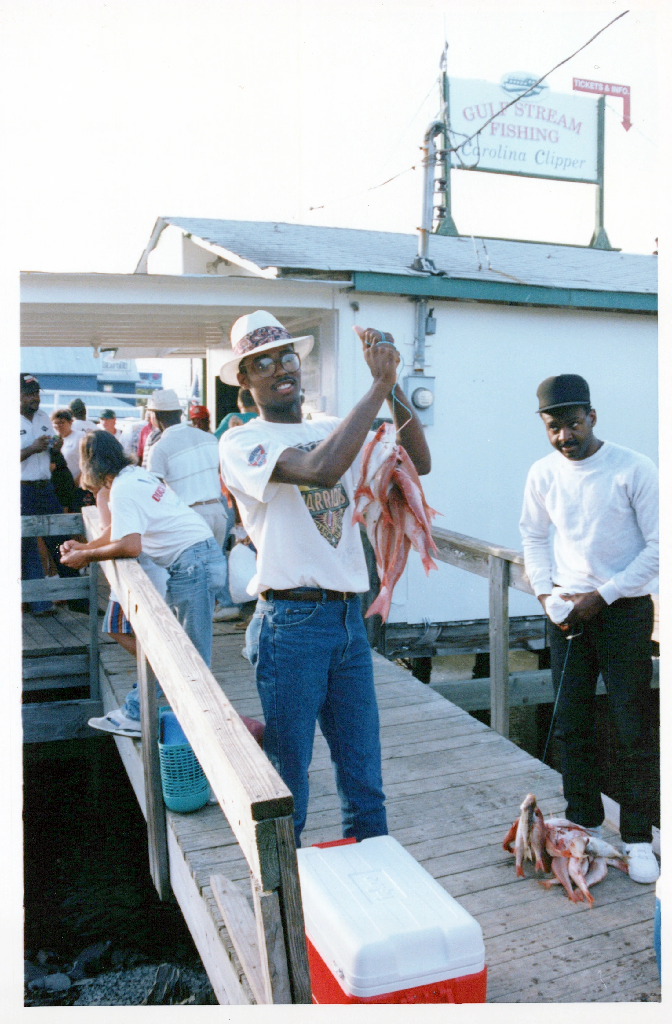 A Black man in a white hat and glasses smiles as he holds up a string of red fish on a wooden dock. Another man and a cooler are nearby.