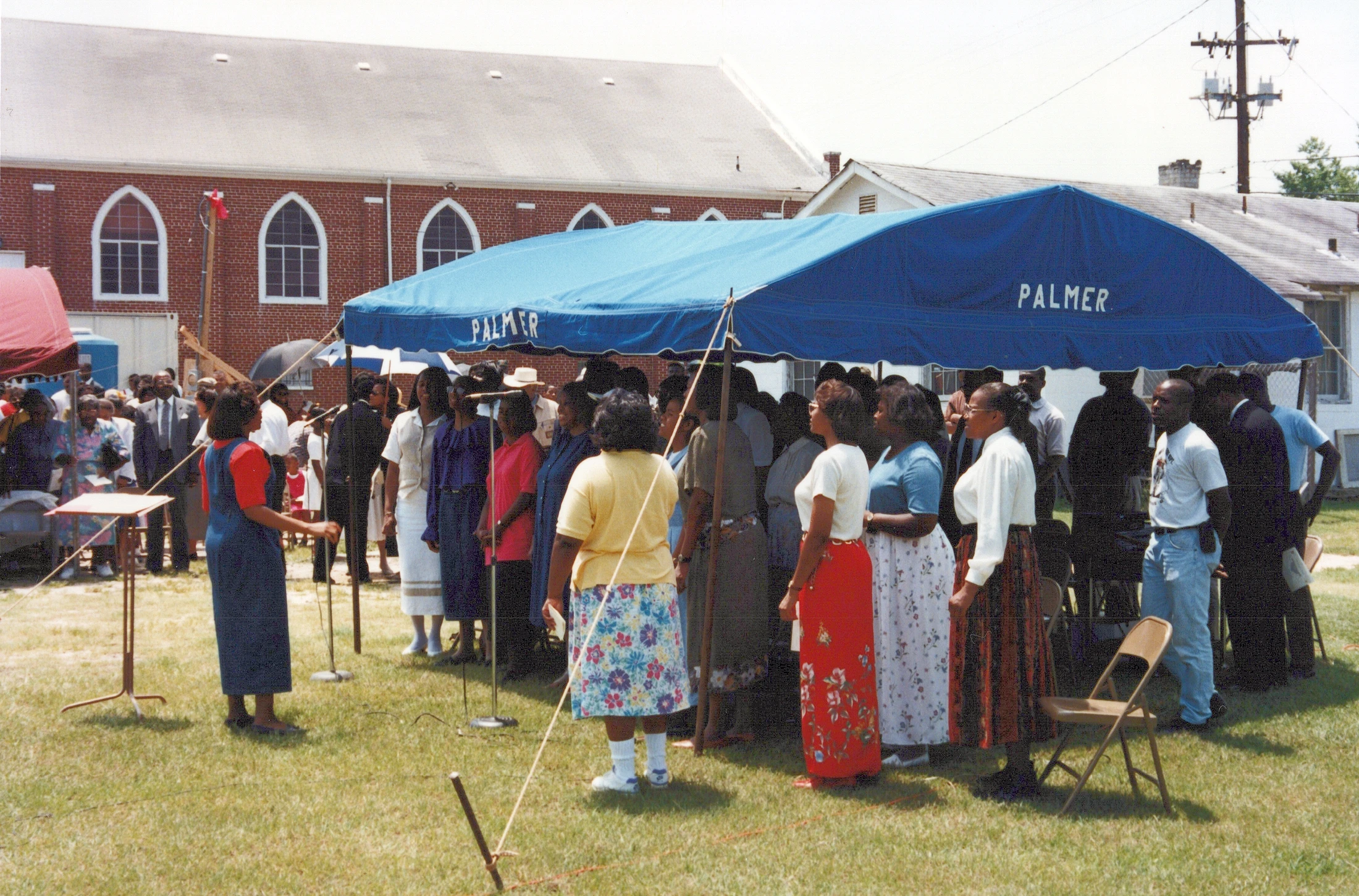 A large group of Black church members gathers on a grassy lawn under a large blue tent. A woman in a blue outfit speaks at a lectern in front of them.