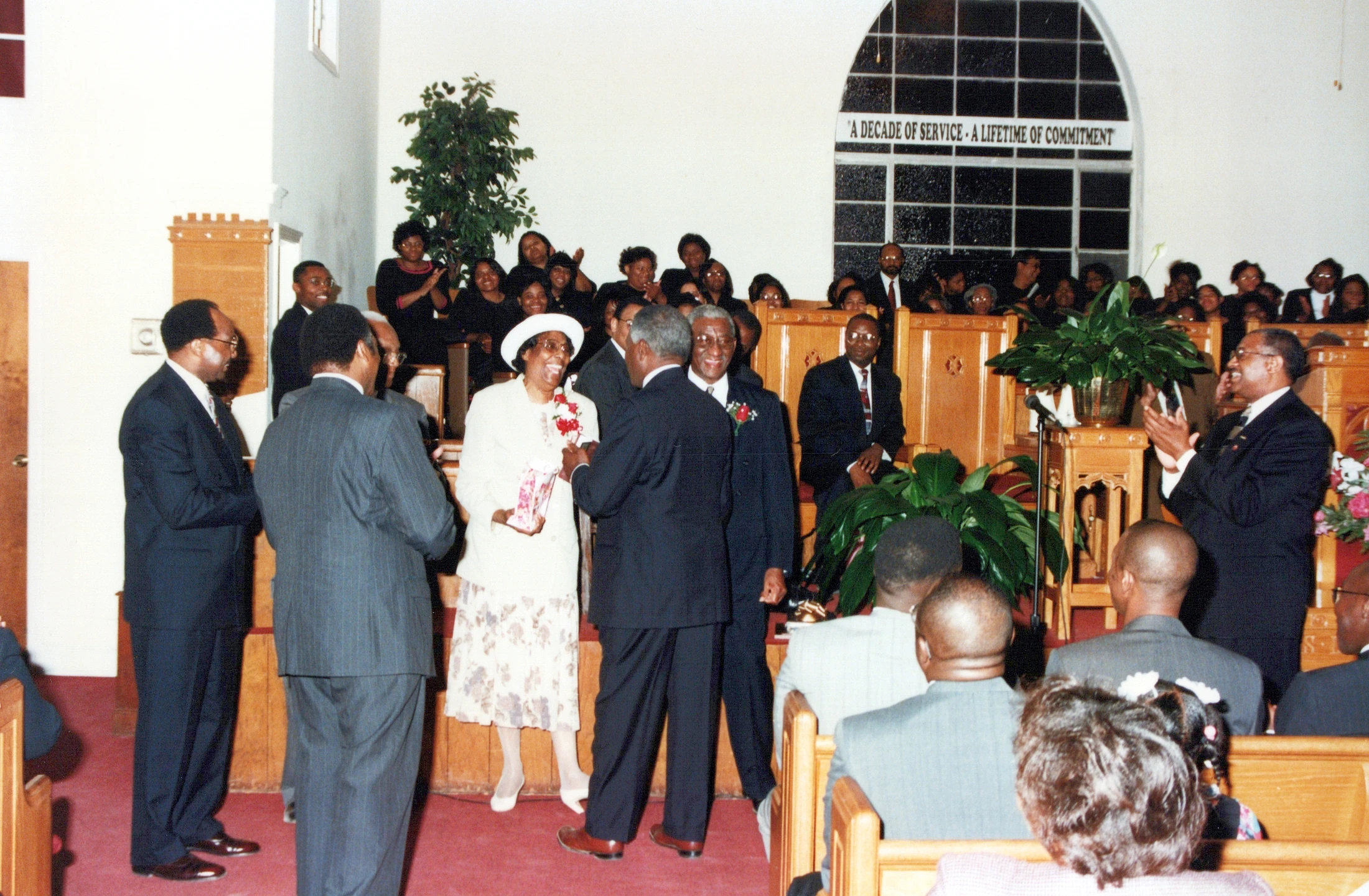 A Black congregation gathers at the front of a church to celebrate a smiling woman in a white suit and hat, honoring her for a decade of service.