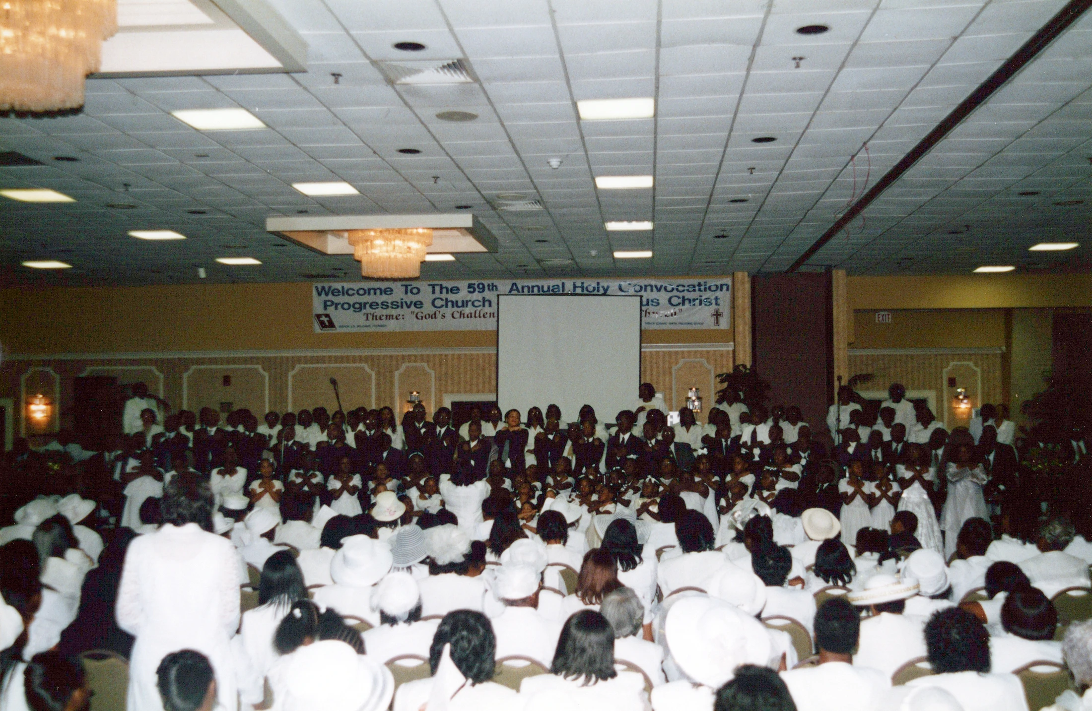 A large congregation, mostly dressed in white, fills a hall for the 59th Annual Holy Convocation, facing a stage with a banner.