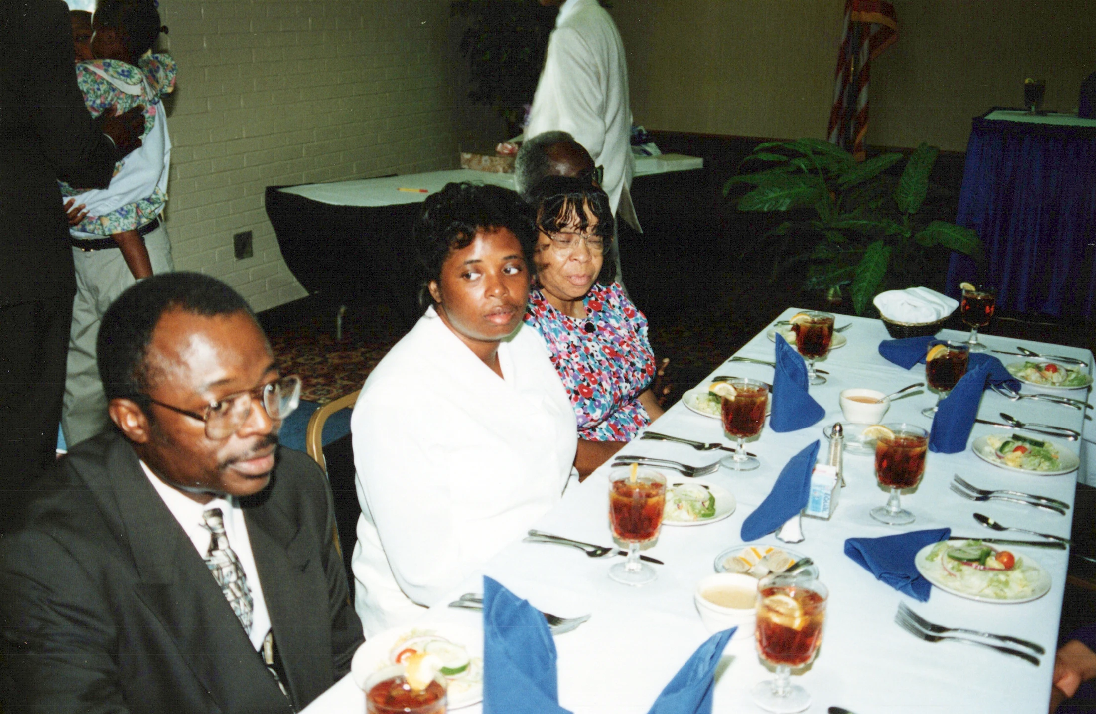 A man in a suit and two women are seated at a long, formally set dinner table with salads and drinks during an indoor event.