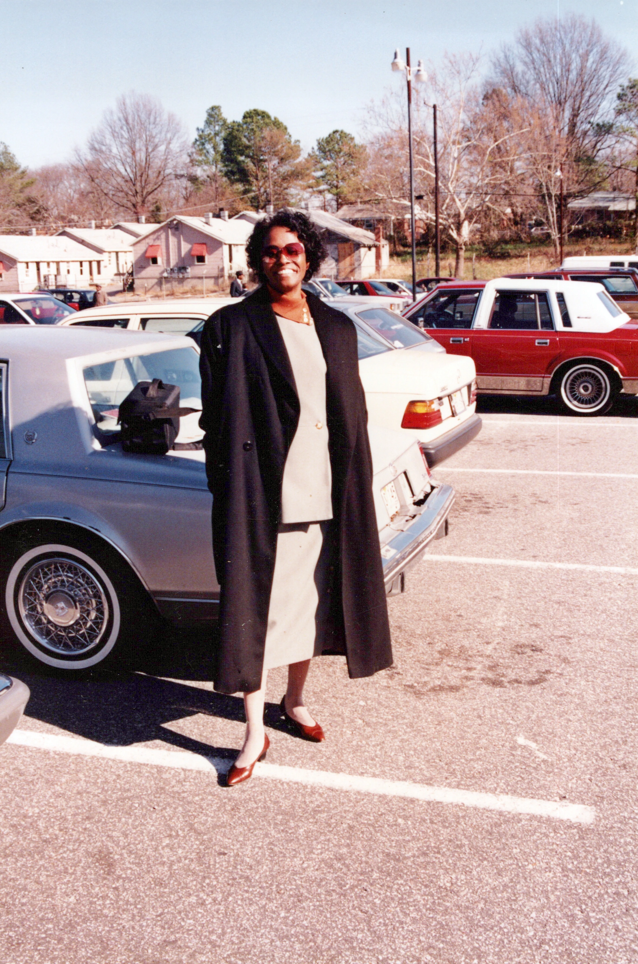 A vintage color photo of a smiling Black woman wearing a long black coat and sunglasses, posing next to a car in a sunny parking lot.