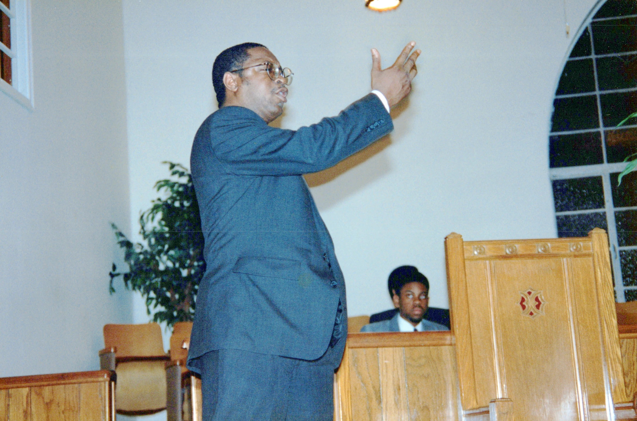 A Black man wearing a dark suit and glasses stands speaking in a church, with his hand raised in a passionate gesture. Another man sits in a pew behind him.