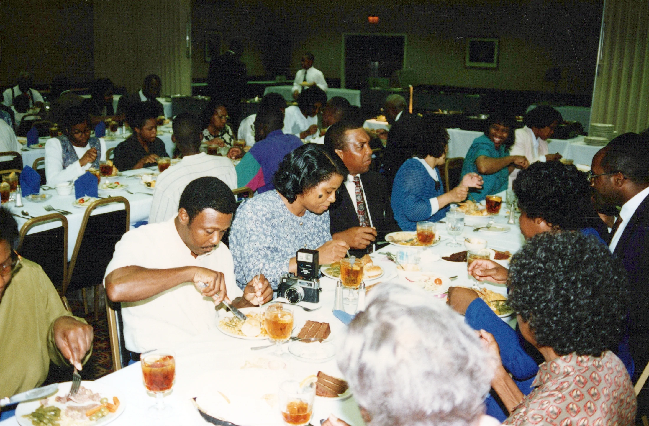A vintage photograph shows a large group of Black men and women eating a meal together at long tables with white tablecloths in a dimly lit banquet hall.