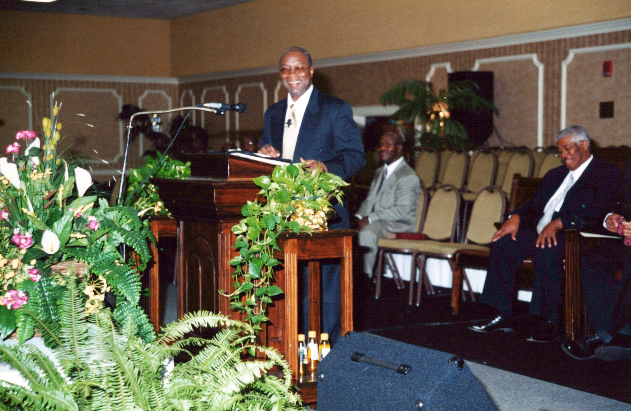 An older Black man in a dark suit and glasses smiles while standing at a wooden podium. Two other men in suits sit behind him in a conference hall.
