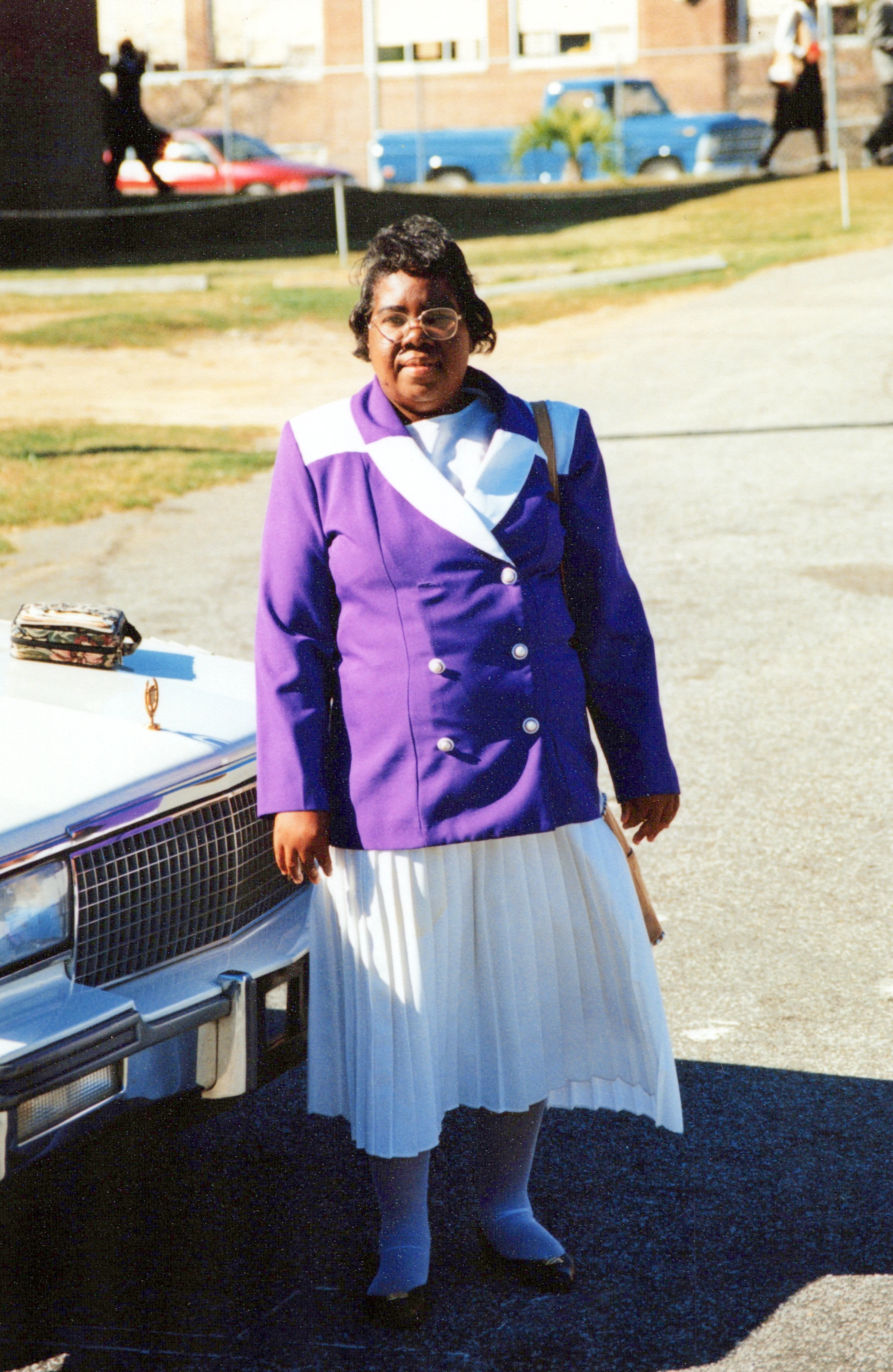 A vintage photo of an older Black woman with glasses standing in a parking lot. She wears a purple and white double-breasted jacket and a white pleated skirt.