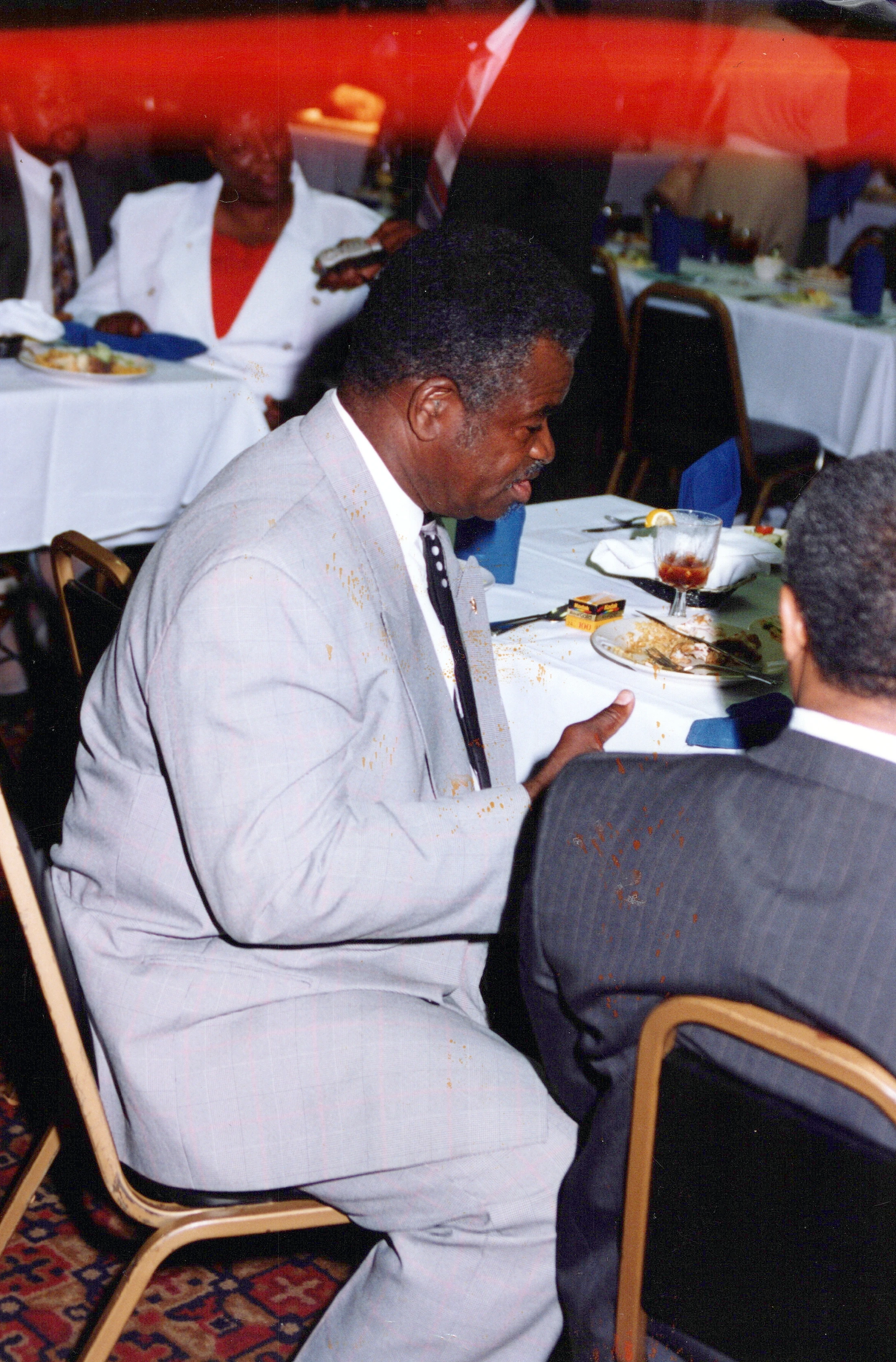A candid photo of a Black man in a light grey suit sitting at a dinner table, turned to his right to talk to another person at a formal event.