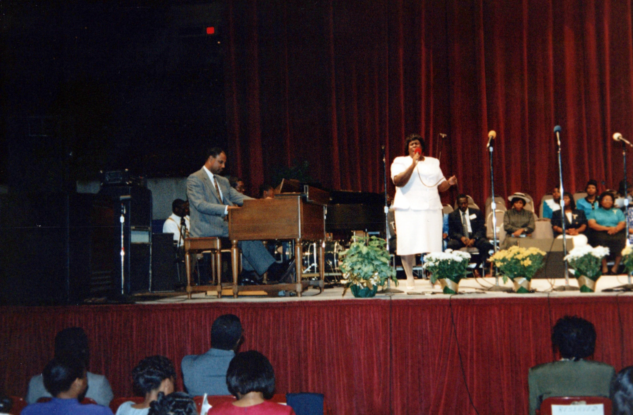 A Black woman in a white suit sings into a microphone on stage, accompanied by a man playing the organ. An audience watches from below.