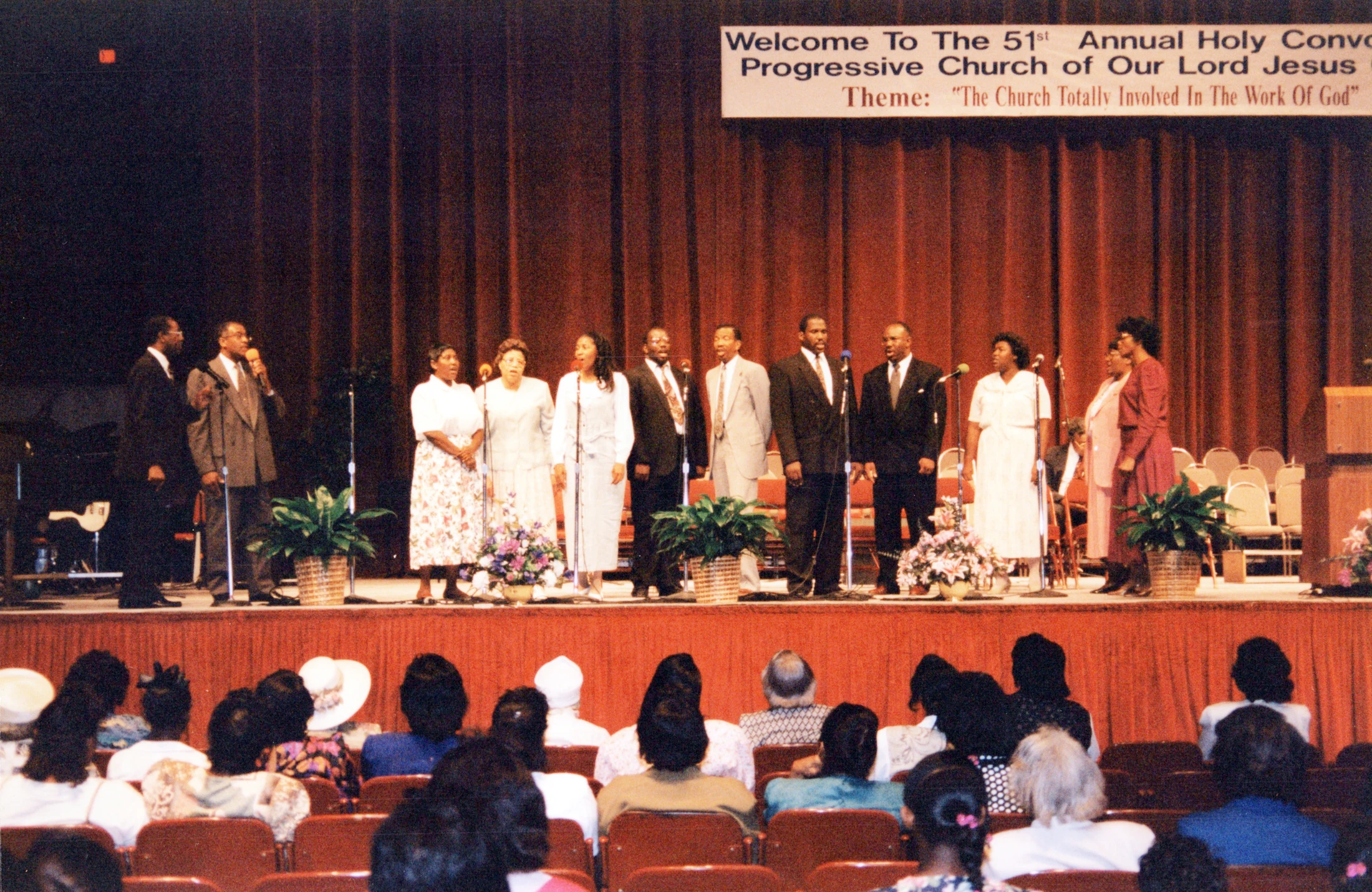 A choir of Black men and women perform on a stage with a banner for the '51st Annual Holy Convocation' behind them as an audience watches from red seats.