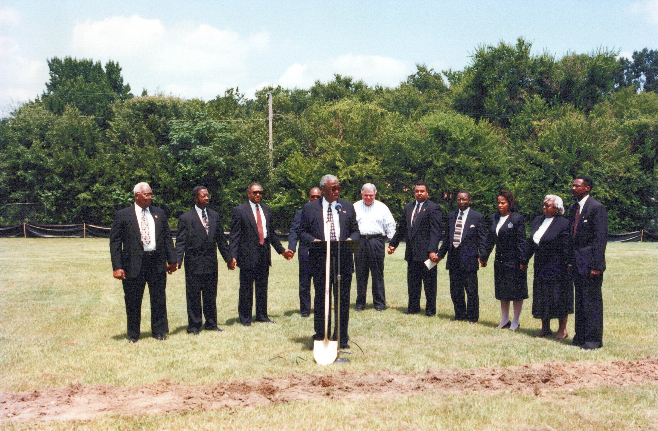 A diverse group of people in suits hold hands in a grassy field for a groundbreaking ceremony. A man stands at a podium with a shovel in front of him.