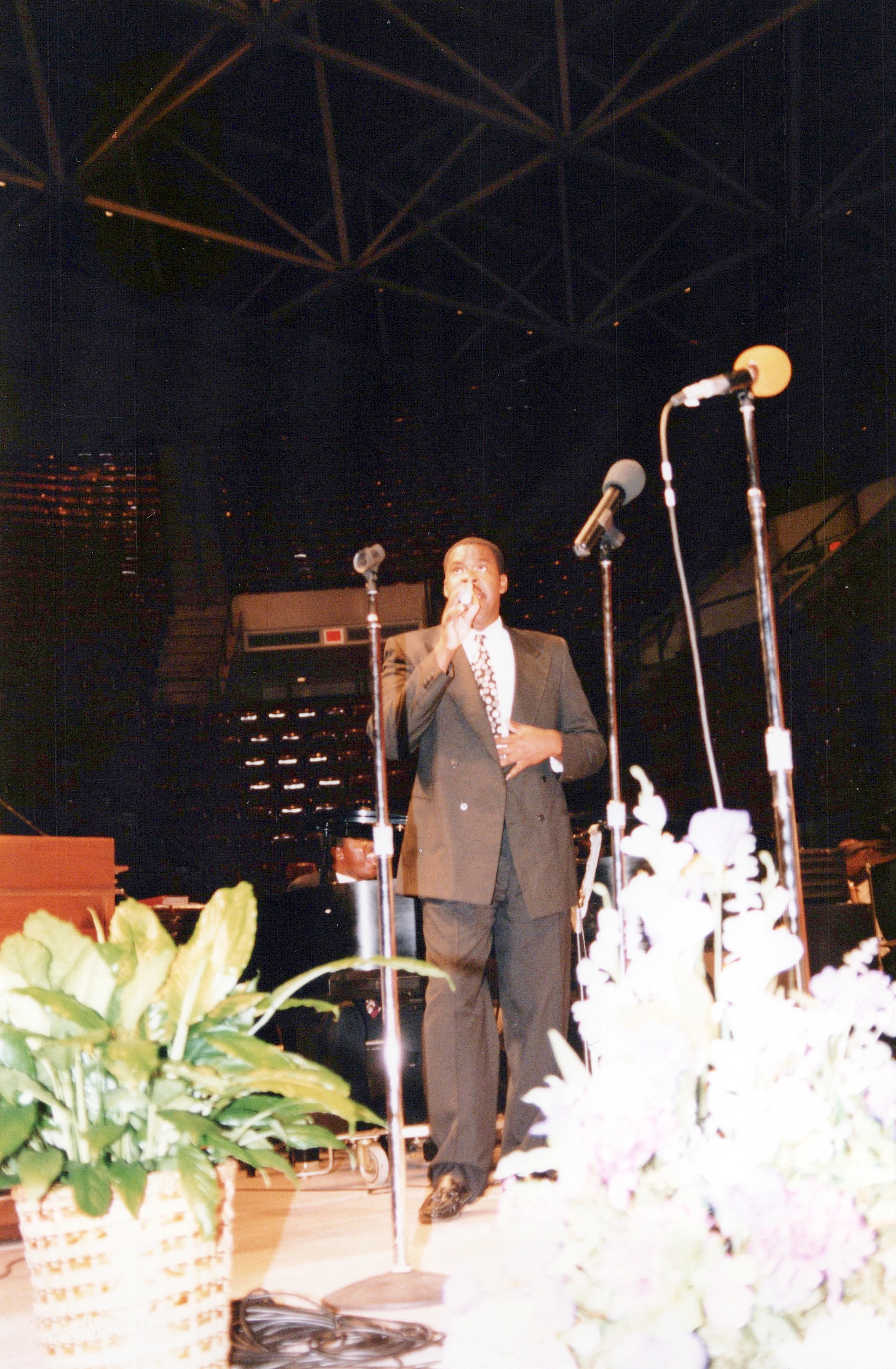 A Black man in a dark suit stands on a large stage, singing into a microphone. The foreground has blurry plants, and the background shows a dark auditorium.