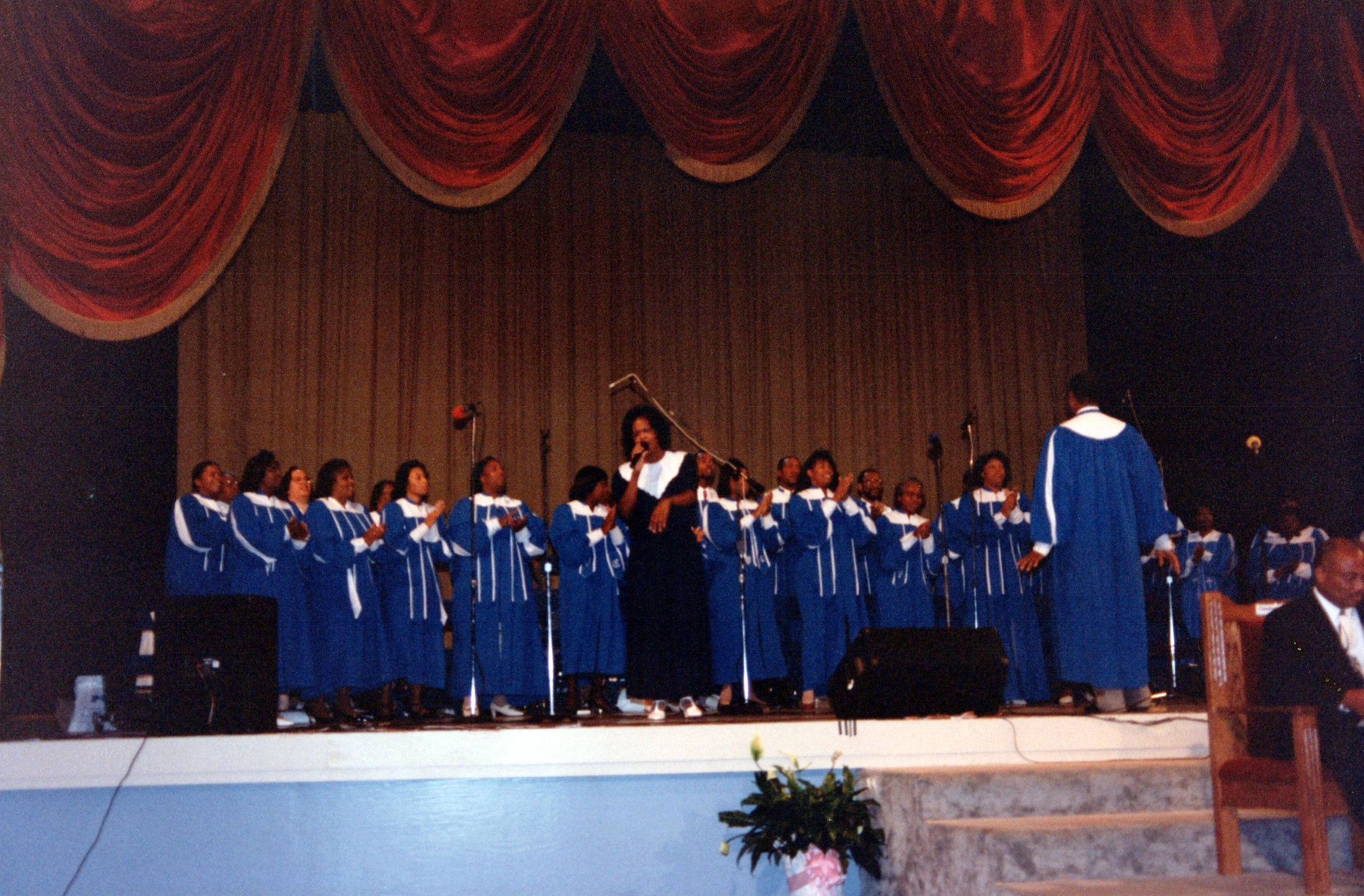 A vintage photograph of an African American gospel choir in blue robes singing on a stage with a large red curtain backdrop.