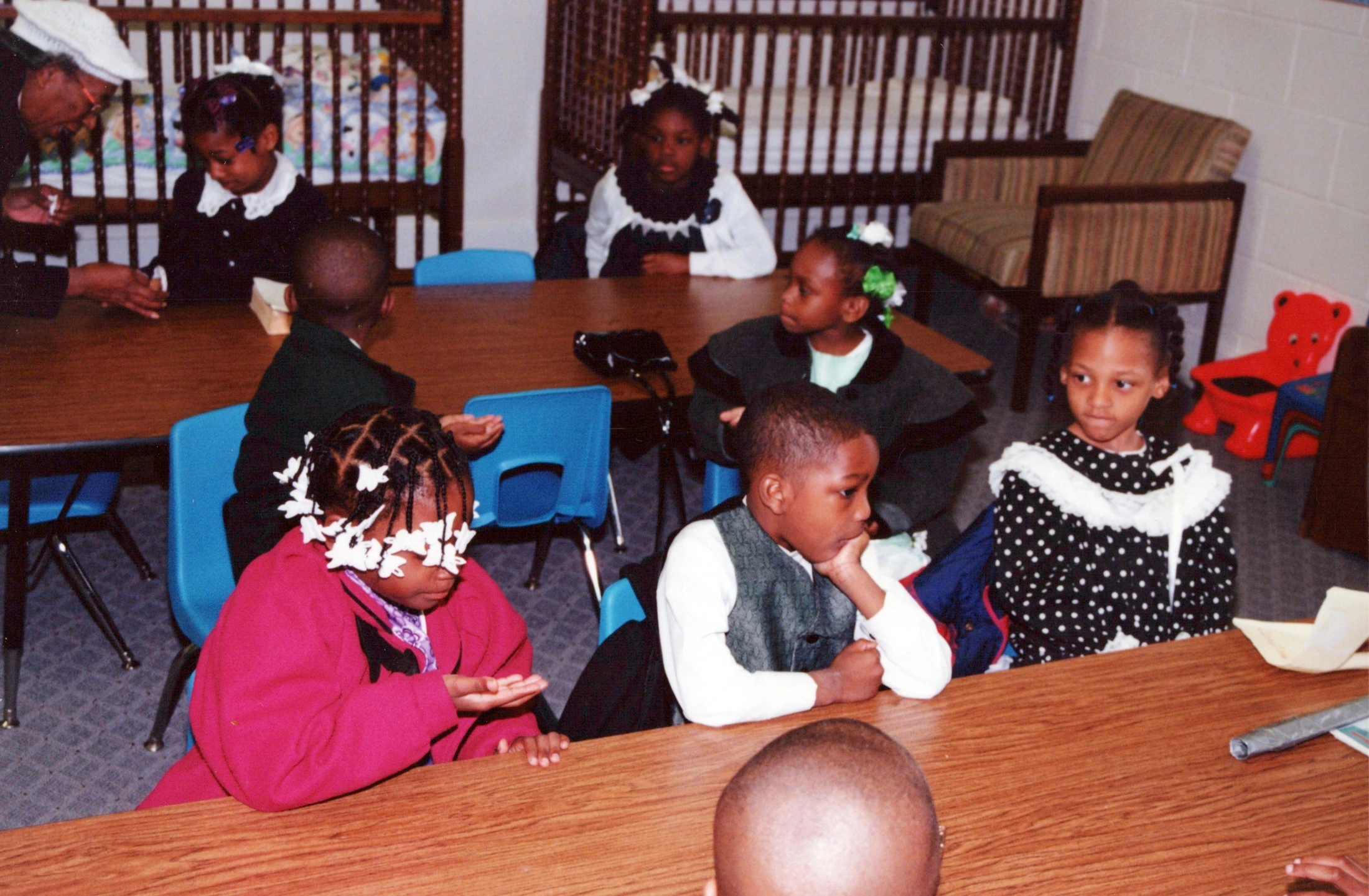 A group of young African American children dressed up, sitting at wooden tables in a classroom or daycare, with an adult interacting with one child.