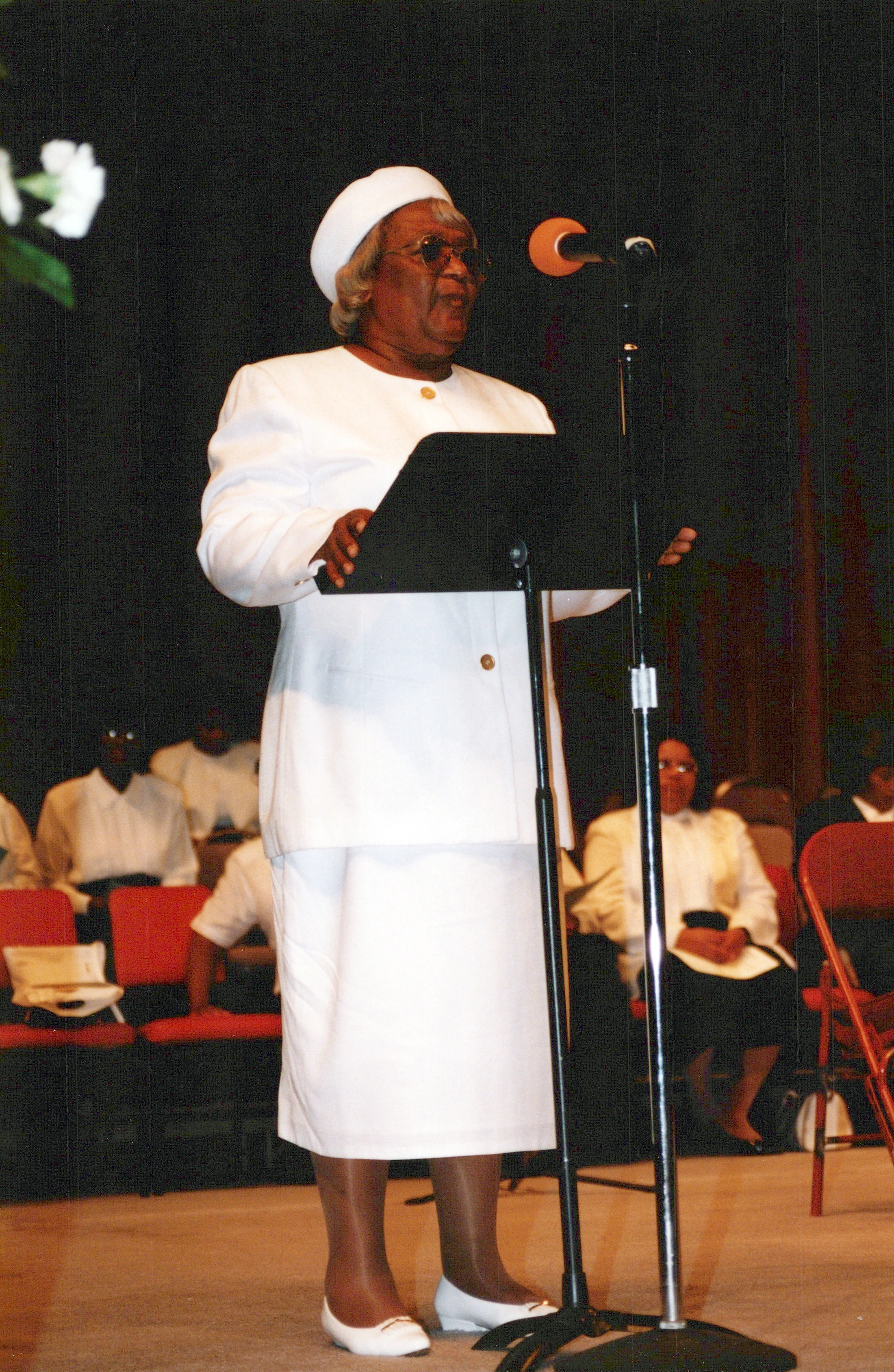 An elderly Black woman wearing a white suit, hat, and glasses stands on a stage at a lectern, addressing an audience seated in the background.