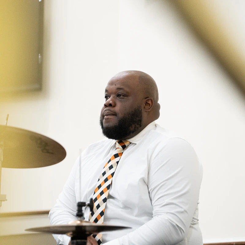 A bald Black man with a beard, wearing a white shirt and a patterned tie, sits thoughtfully behind his drum kit in a brightly lit room.