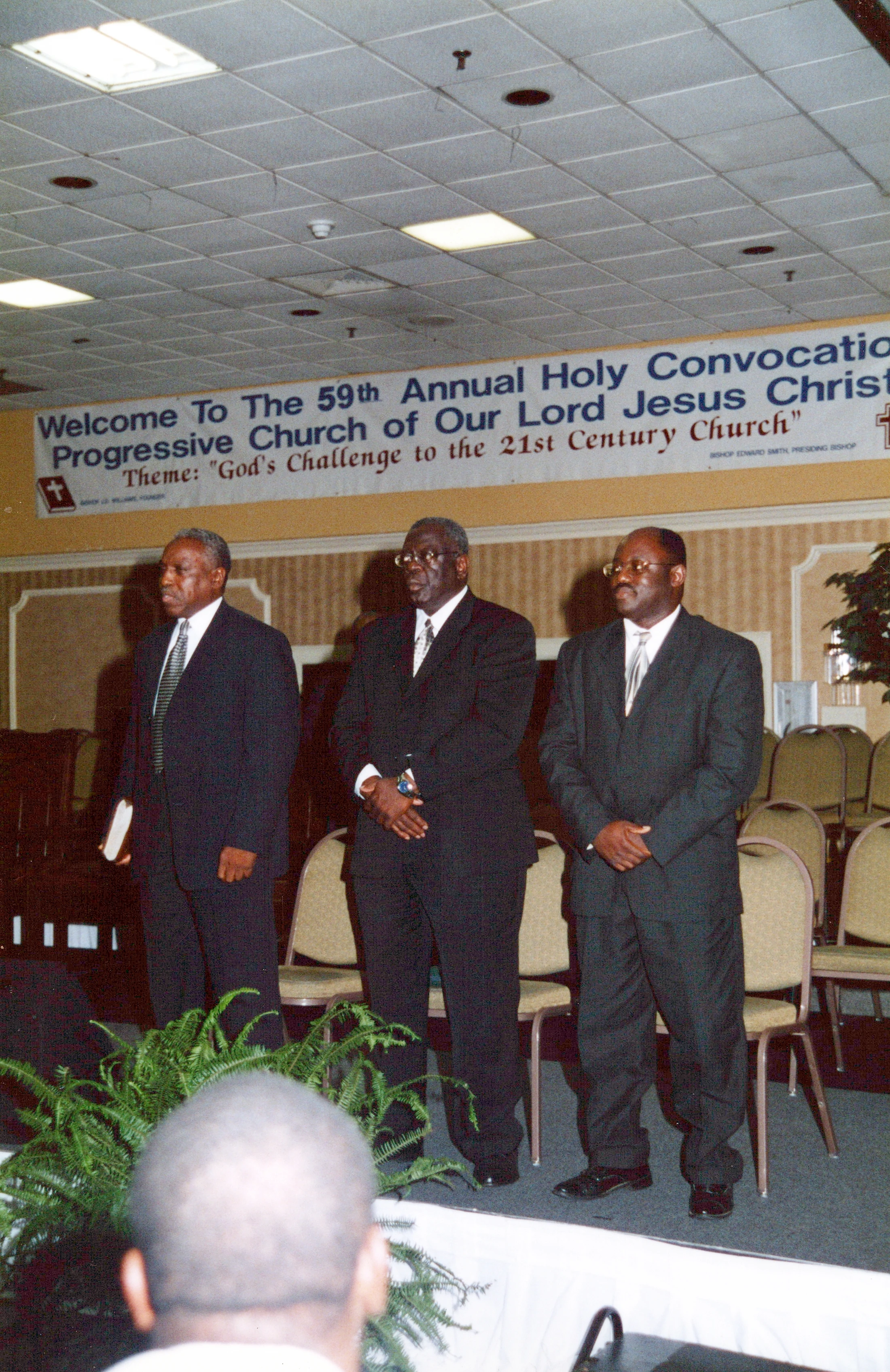 Three Black men in dark suits stand on a stage in front of a large banner for the "59th Annual Holy Convocation Progressive Church of Our Lord Jesus Christ."