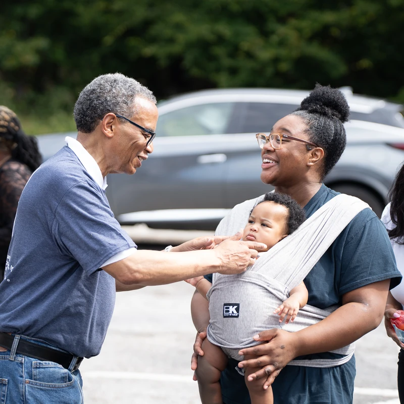 An older Black man with glasses smiles while talking to a young woman holding a baby in a sling. They are all standing outside in a parking lot.