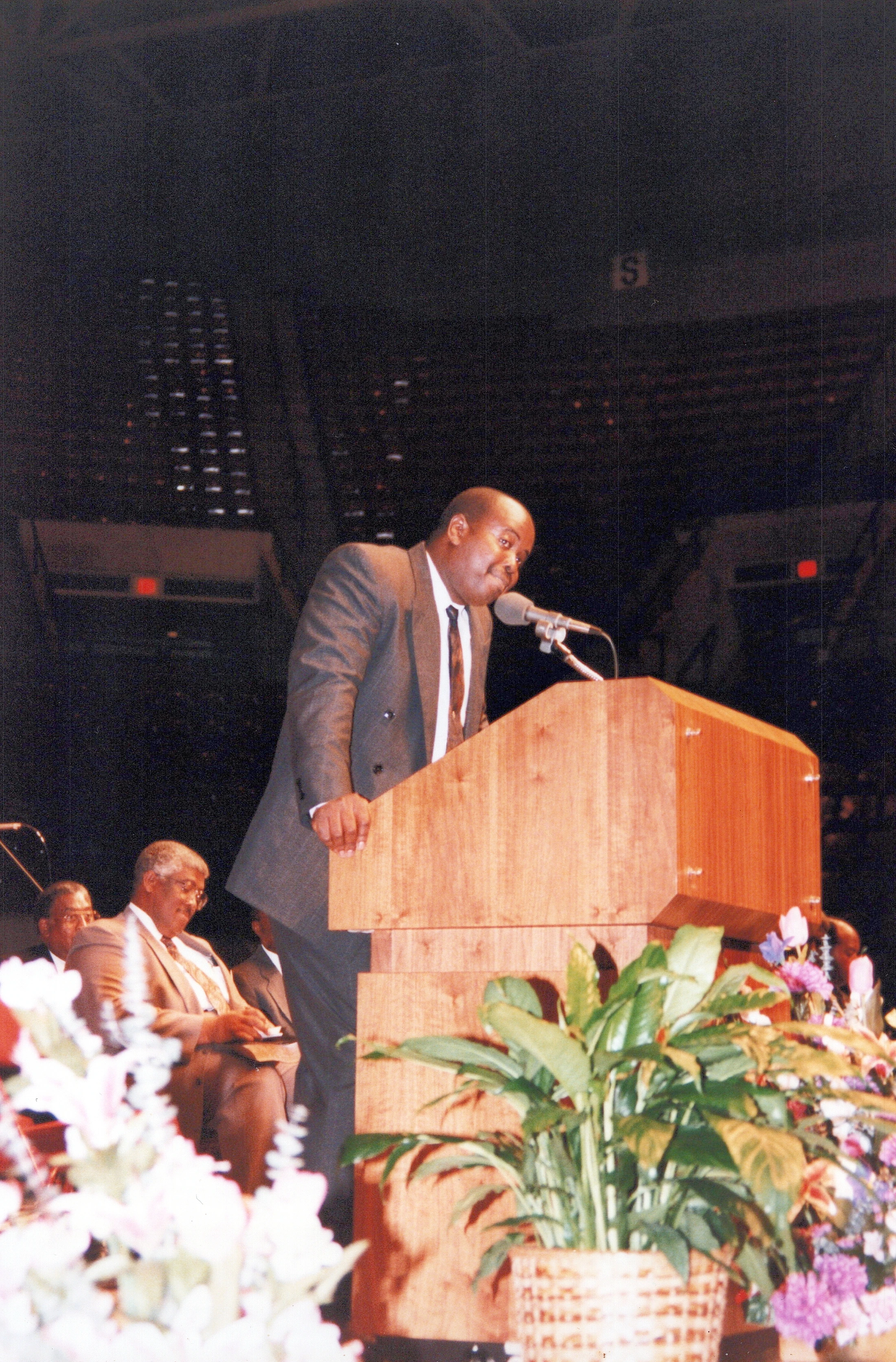 A Black man in a dark suit and tie stands at a wooden lectern, leaning into a microphone to give a speech in a large, dimly lit auditorium.