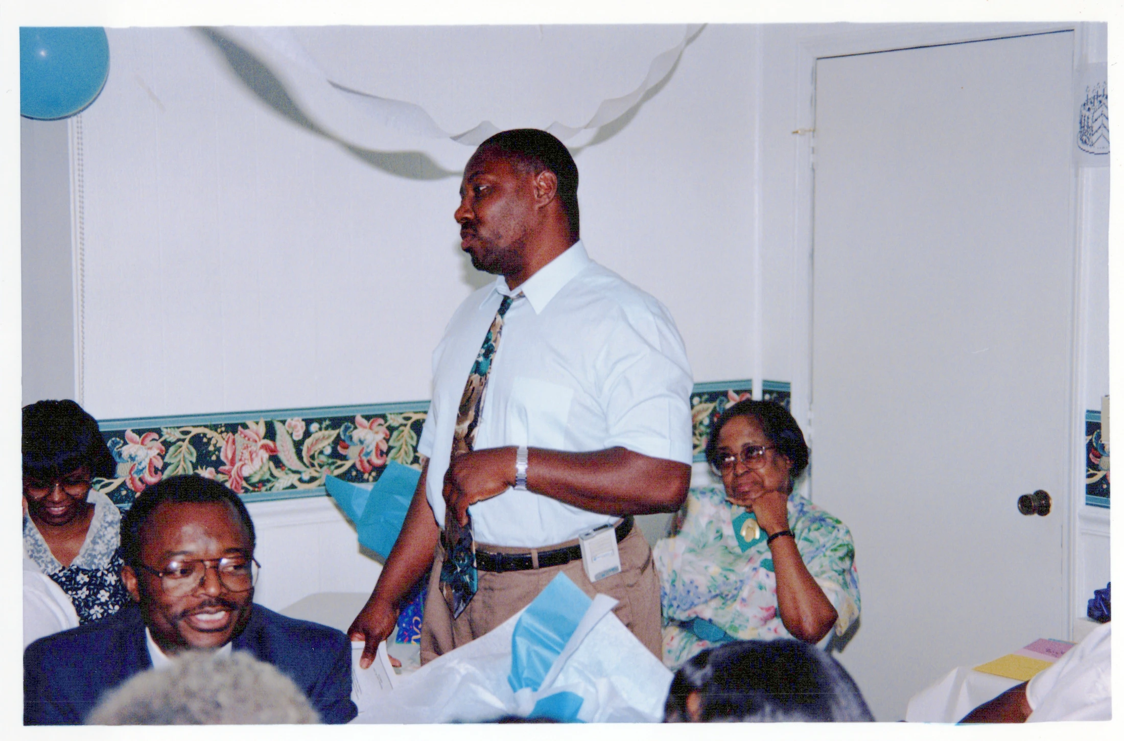A Black man in a shirt and tie stands and speaks at an indoor gathering. Other guests are seated at tables, listening to him.