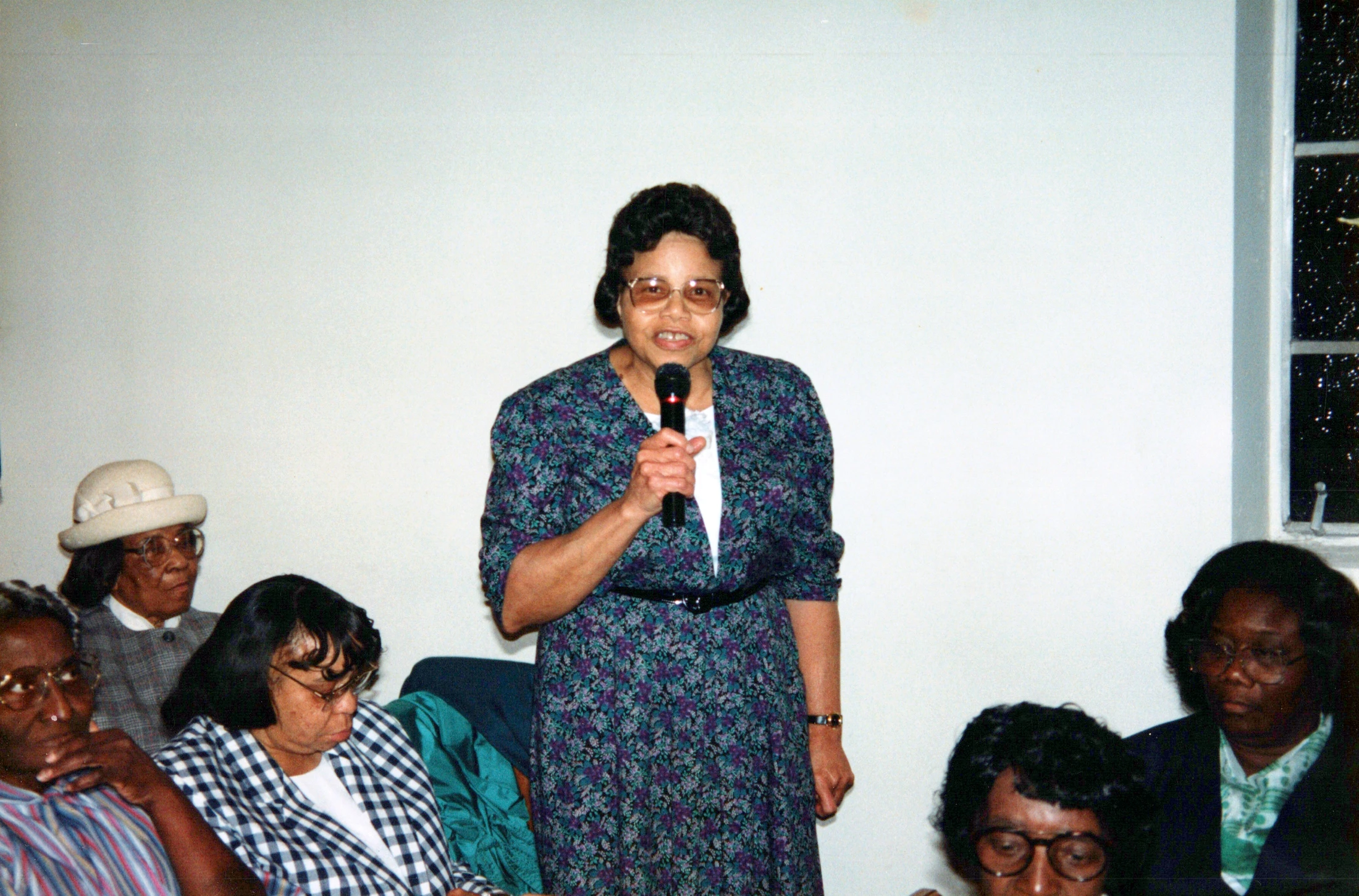 An older Black woman in a floral dress and glasses stands and speaks into a microphone while a group of people sit and listen in an indoor setting.