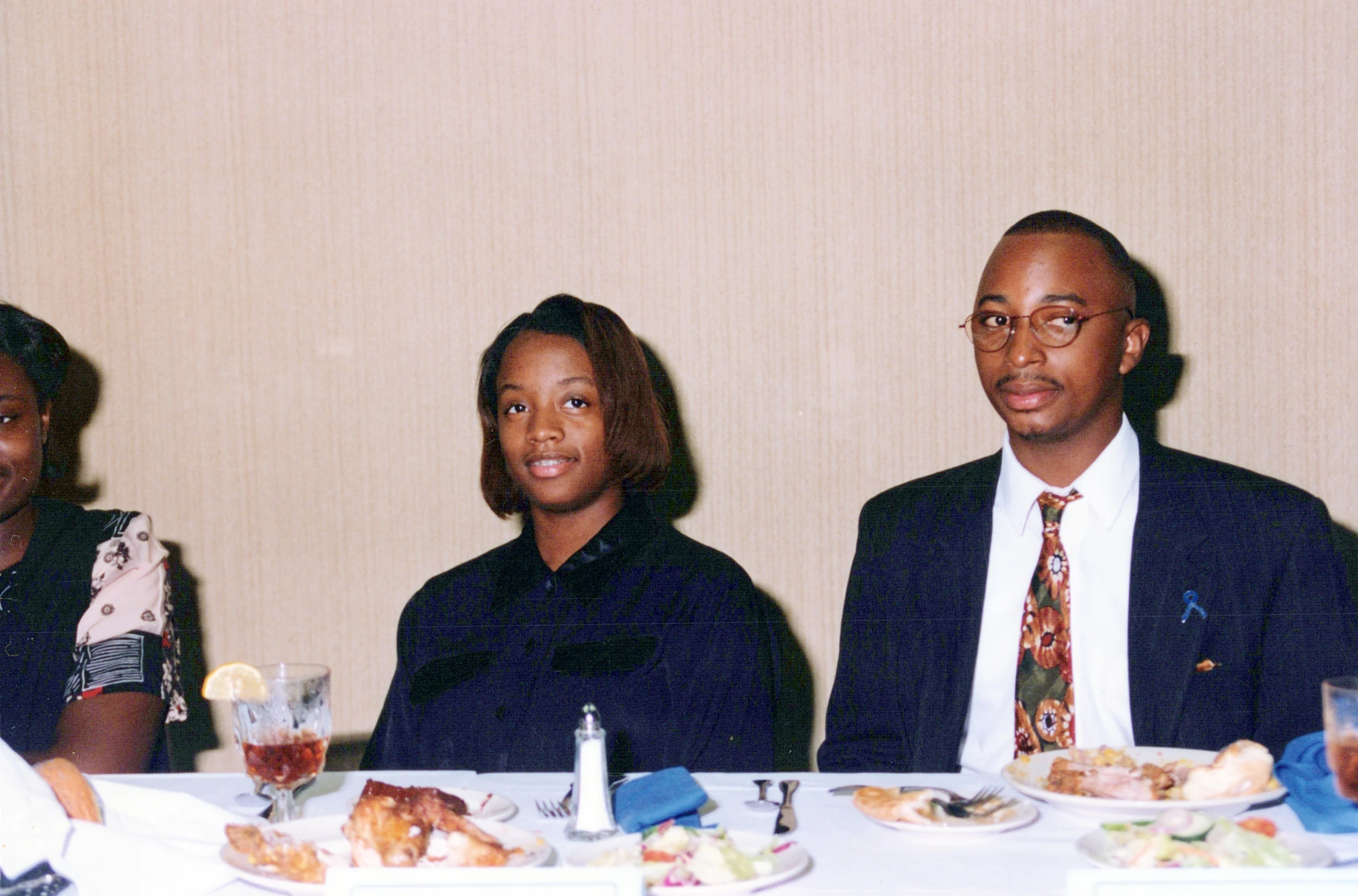 A vintage photograph shows two women and a man, all Black, sitting at a formal dinner table, looking at the camera with plates of food in front of them.