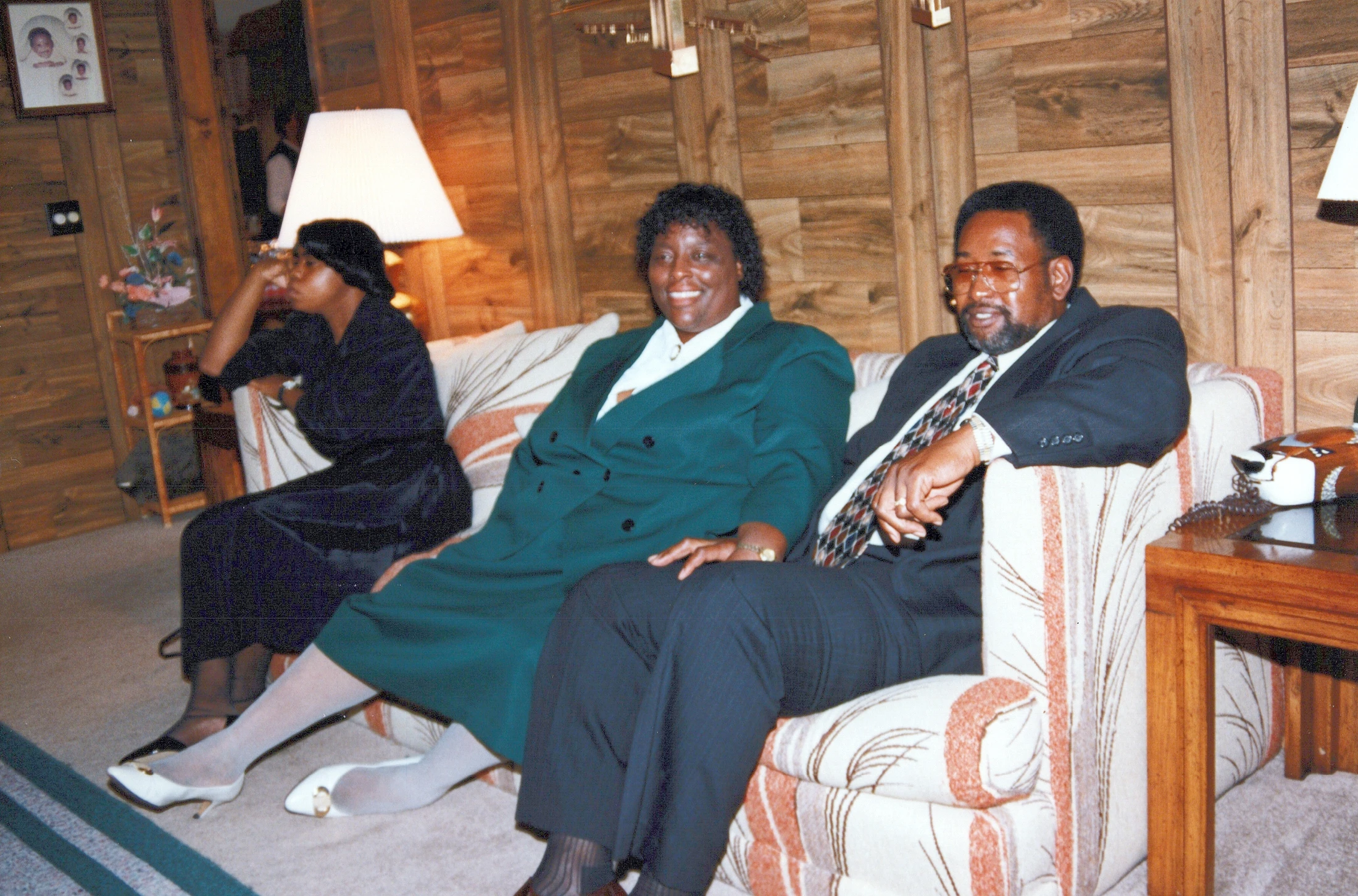 A vintage photograph of three Black adults dressed up and sitting on a patterned couch. A man and a woman in the middle are smiling at the camera.