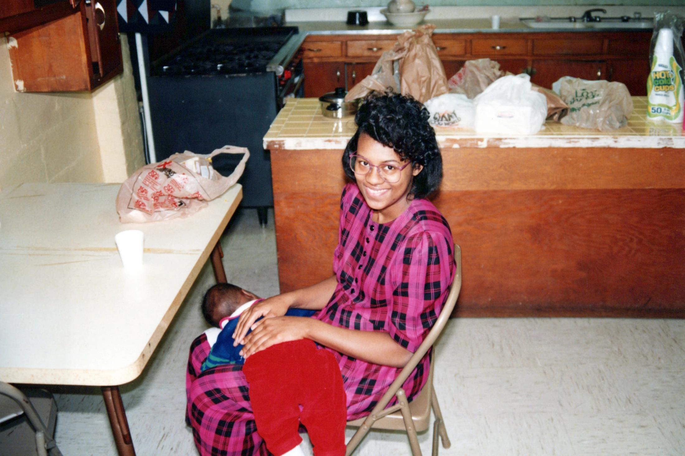 A Black woman with glasses and a pink plaid dress sits on a chair, smiling at the camera while holding a baby in her lap inside a kitchen.