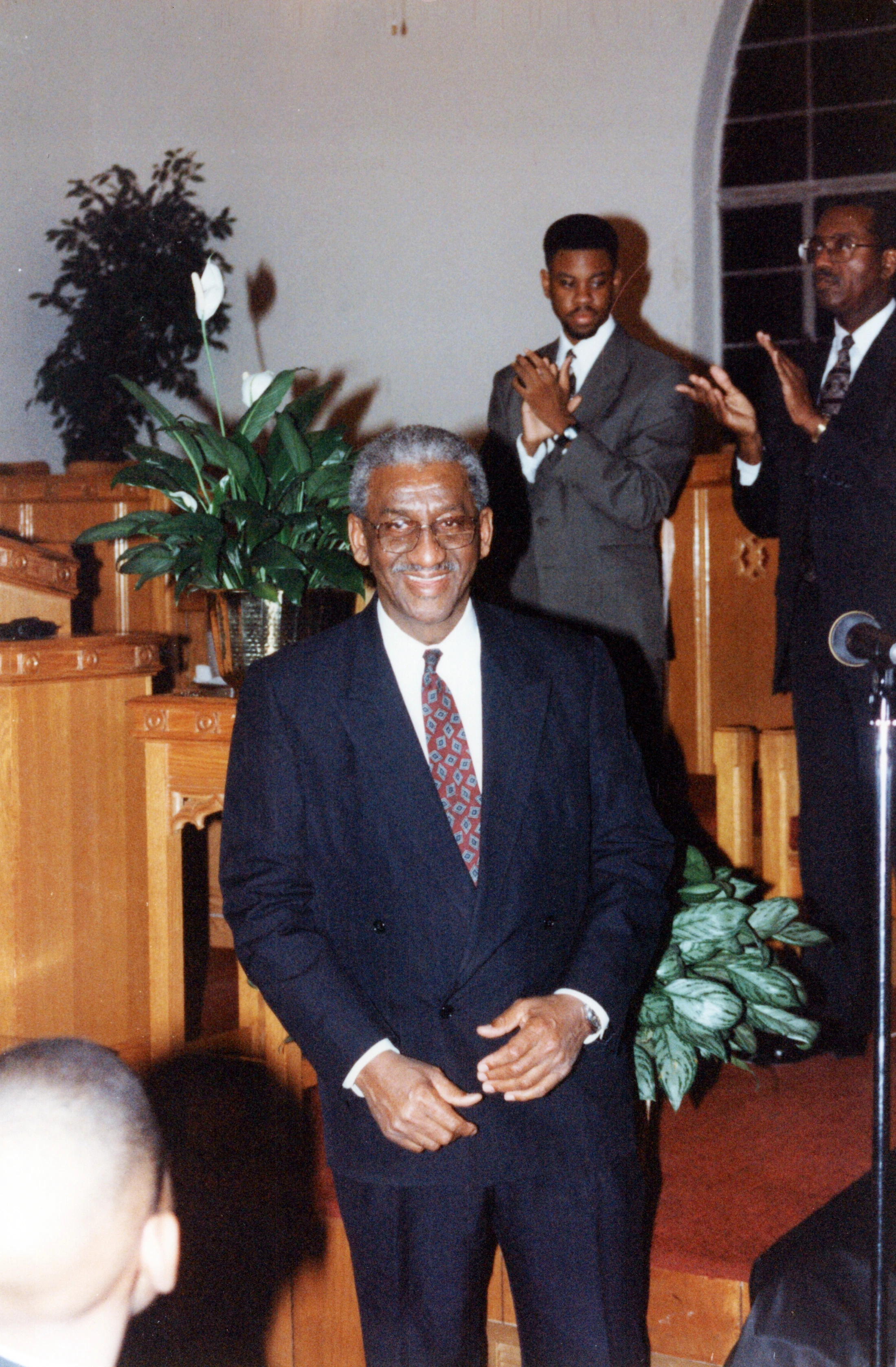 An older Black man with grey hair and glasses smiles in a dark suit. Two younger men stand behind him in a church, clapping and looking on.