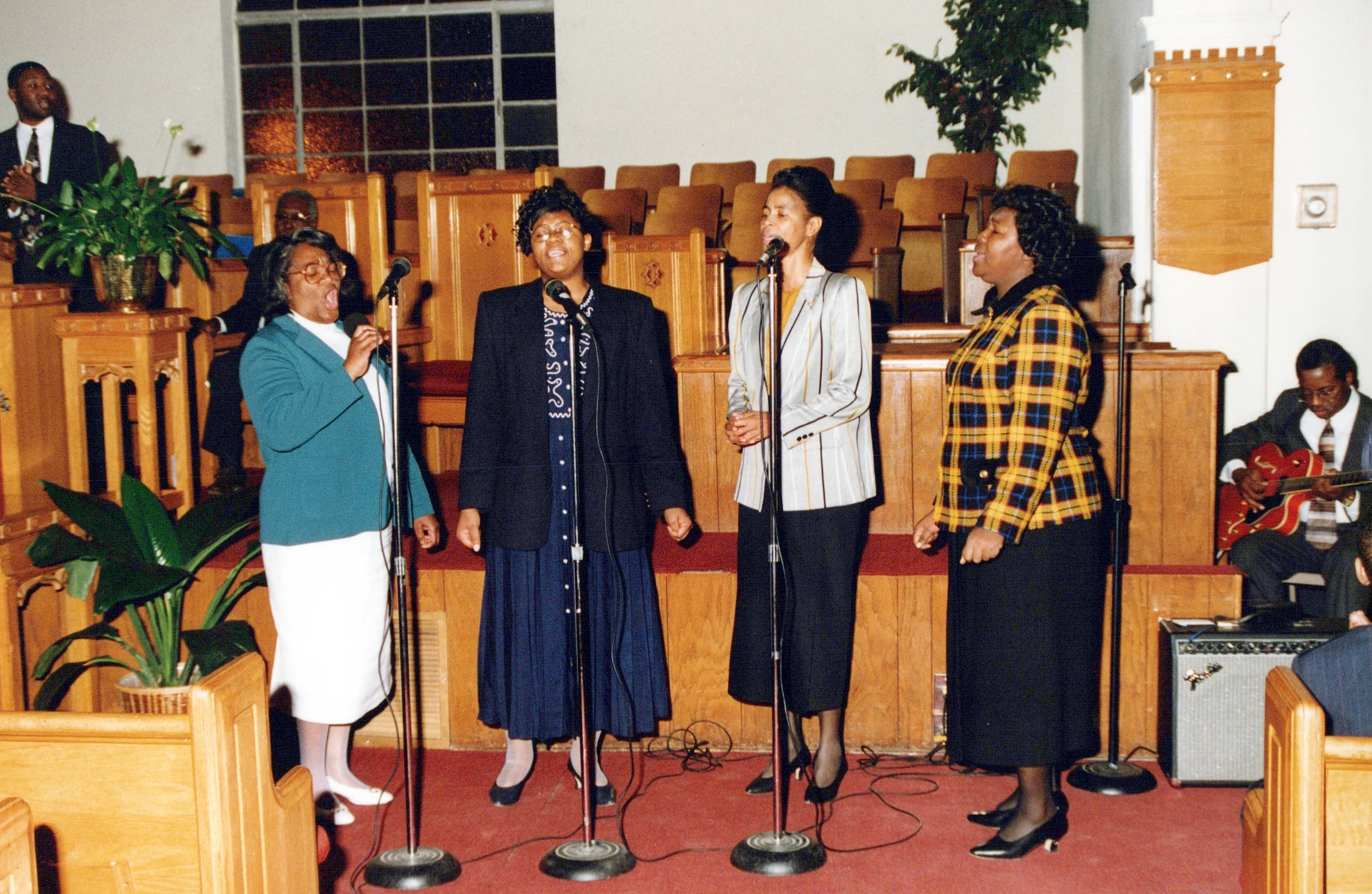 Four Black women in a gospel choir sing passionately into microphones on a stage in a church, accompanied by a man playing an electric guitar.