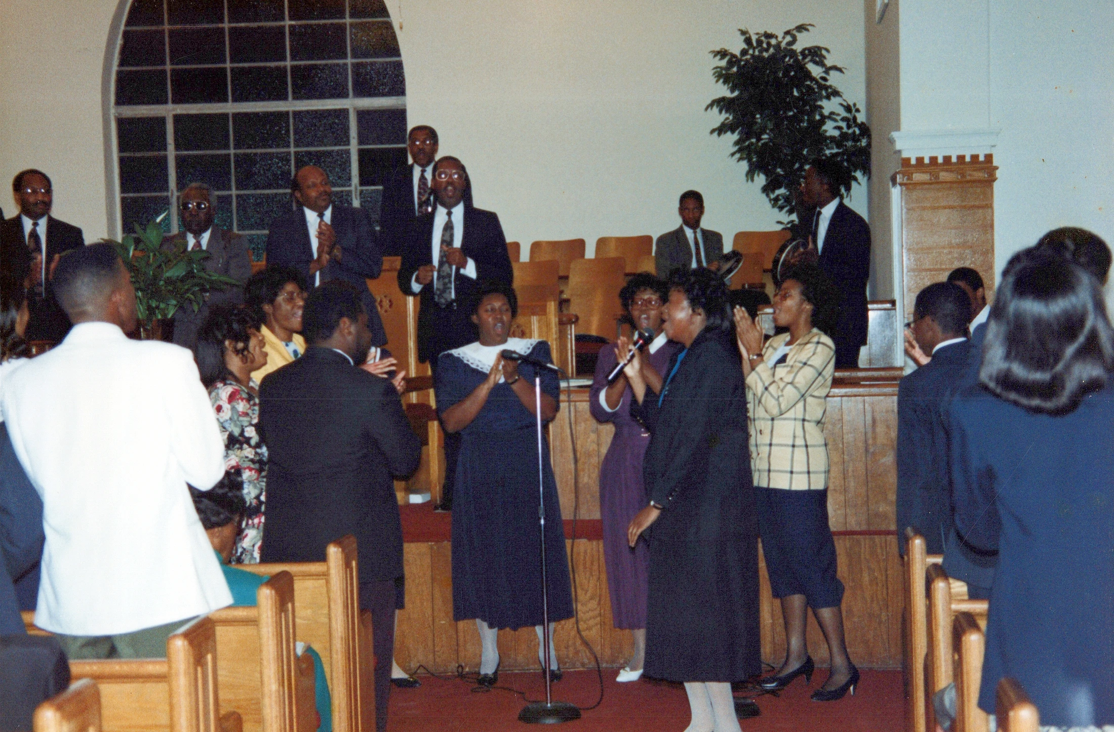 A Black church choir of men and women in suits and dresses sing and clap joyfully at the front of a sanctuary filled with wooden pews.