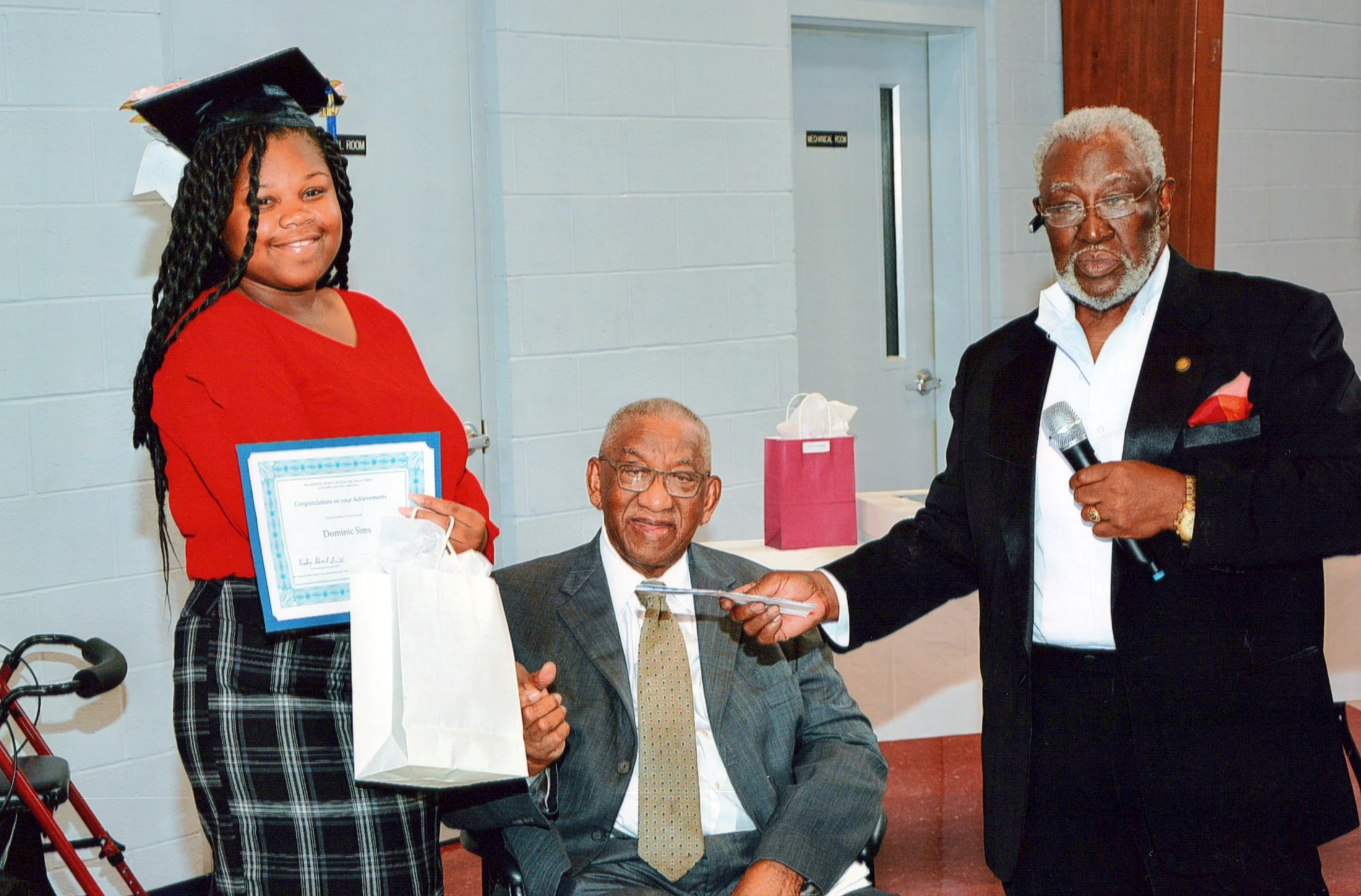 A young Black woman in a graduation cap holds a certificate and gift bag while standing next to two older Black men during a celebration ceremony.