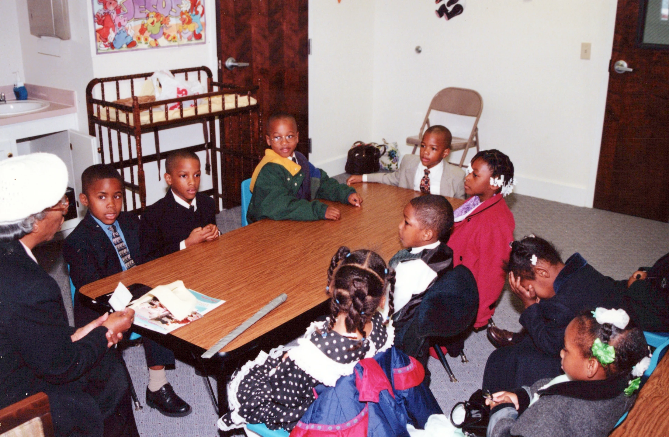 A vintage photograph shows a group of young, well-dressed African American children sitting with an older woman around a wooden table in a classroom.