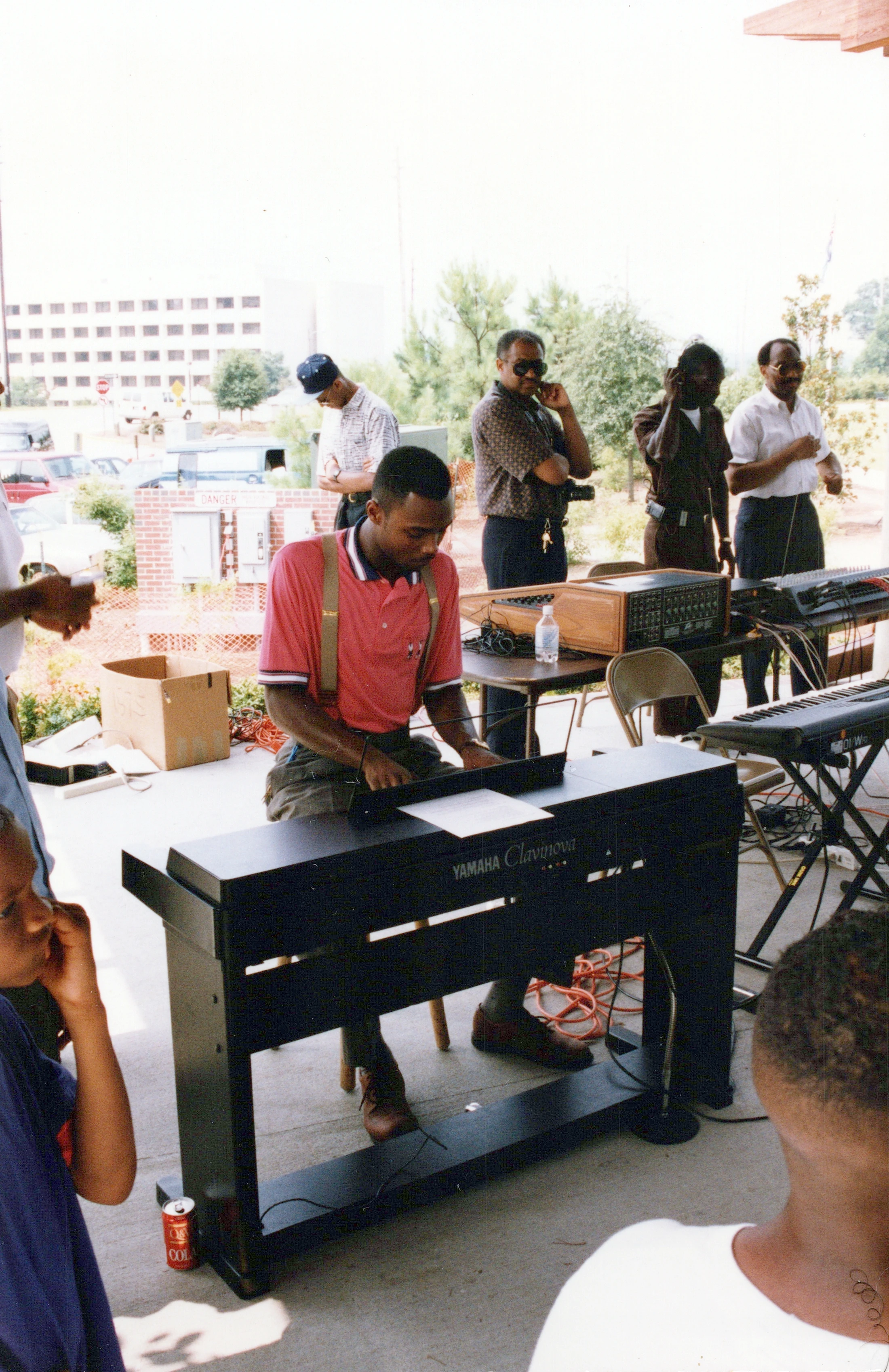 A Black man in a red polo shirt and suspenders plays a Yamaha Clavinova keyboard outdoors on a patio. Other men stand behind him, watching the performance.