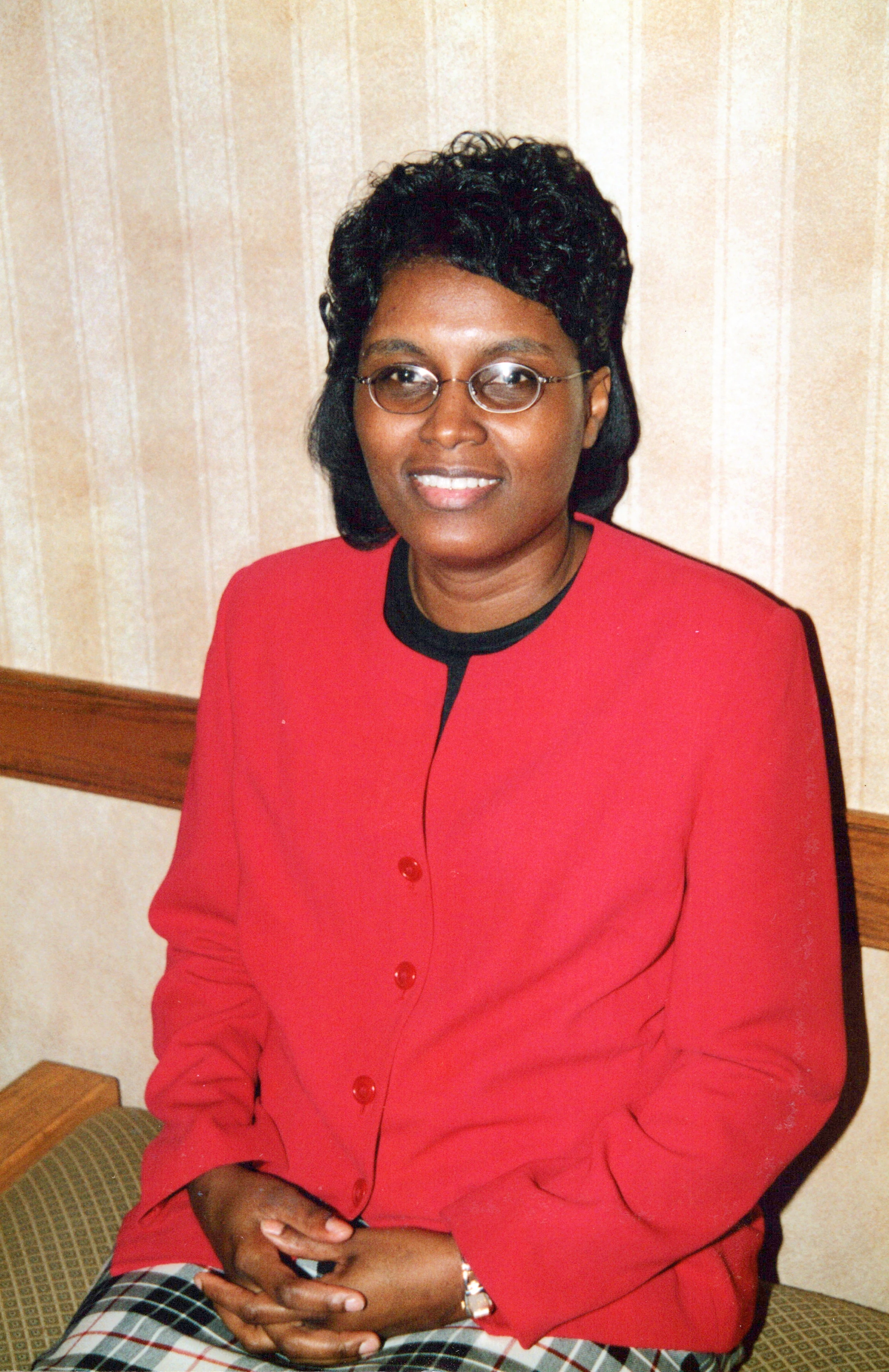 A portrait of a Black woman with short curly hair and glasses, smiling at the camera. She wears a bright red jacket and her hands are clasped in her lap.