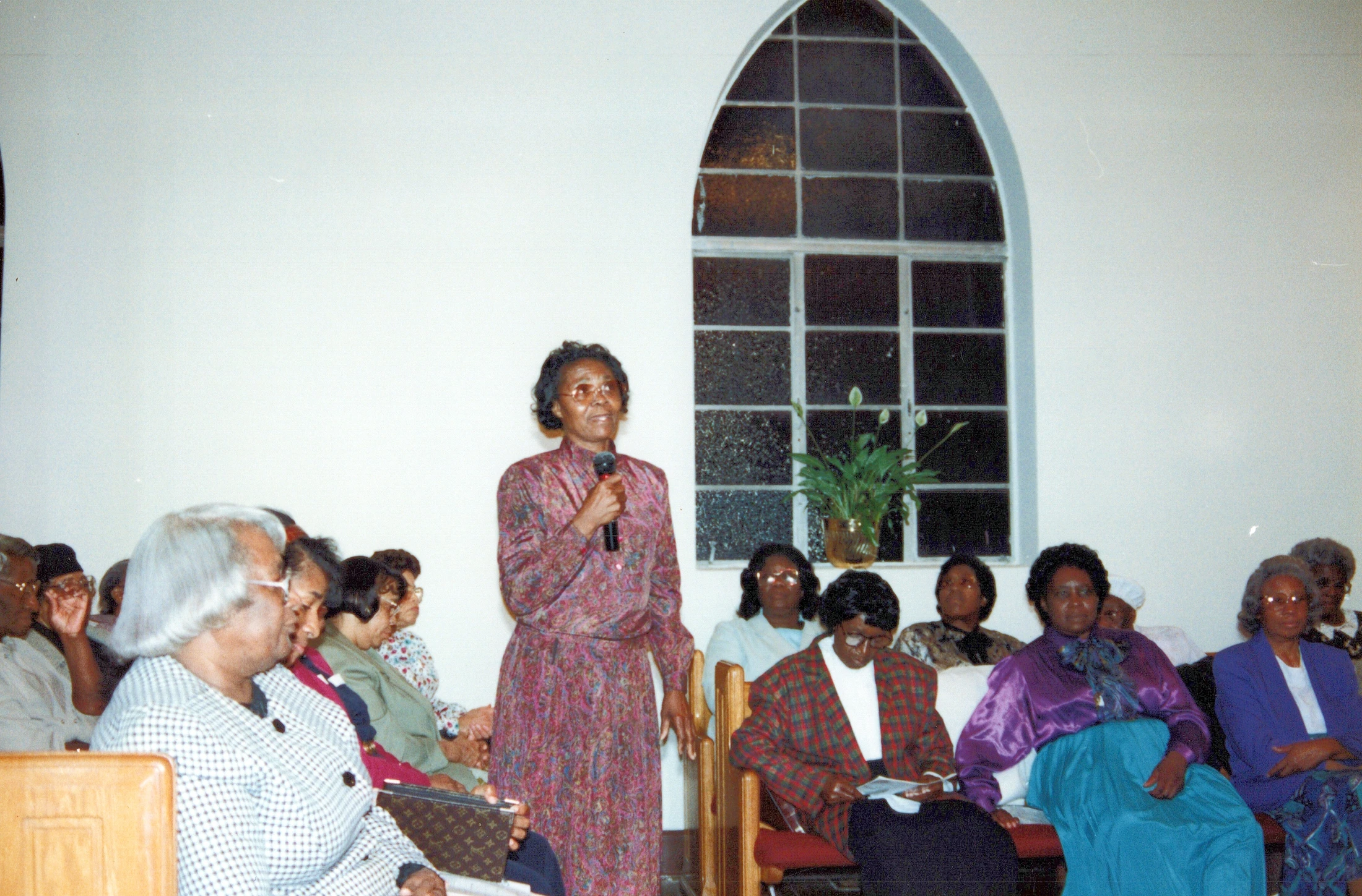 An older Black woman in a patterned dress stands holding a microphone, speaking to a seated audience inside a church with a large arched window.