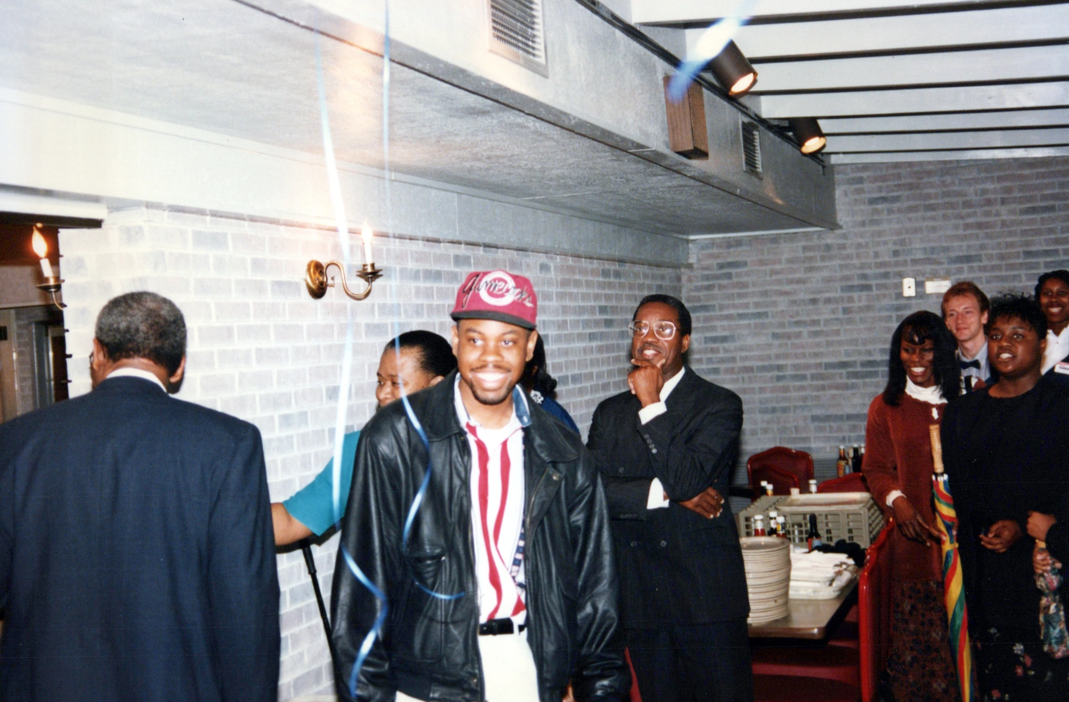 A vintage film photo of a group of people at an indoor gathering. A young man in a leather jacket and red baseball cap smiles in the center.