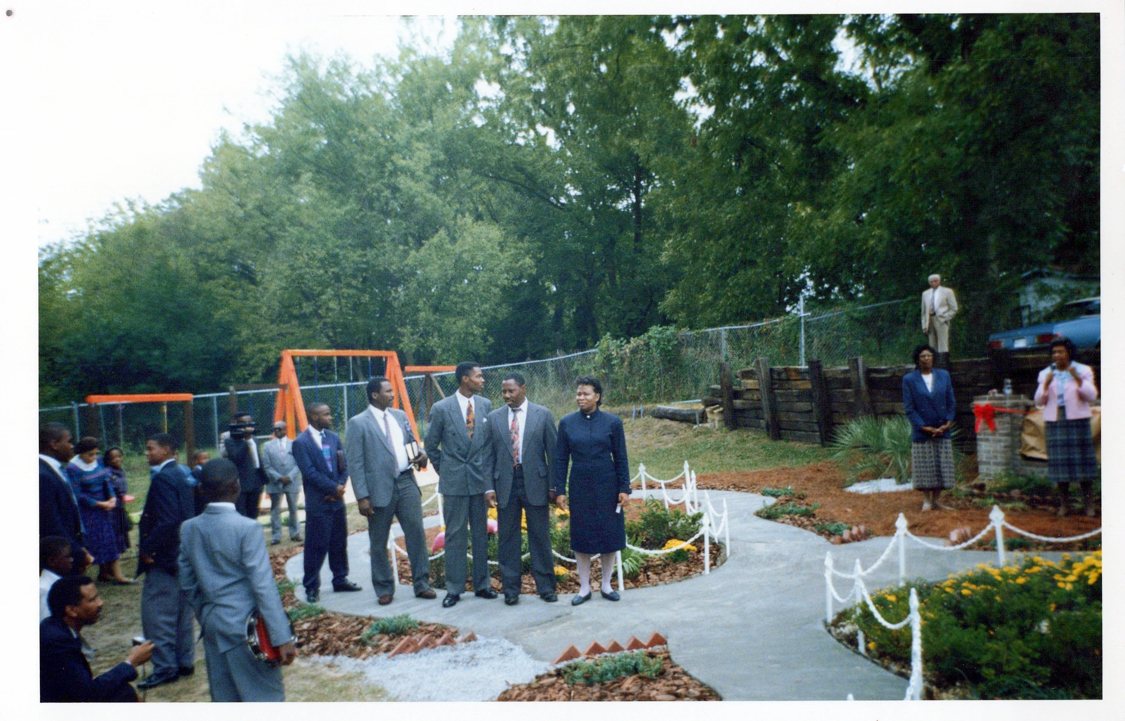 A group of African American men and women in formal attire stand outdoors along a newly landscaped, winding pathway with a playground in the background.