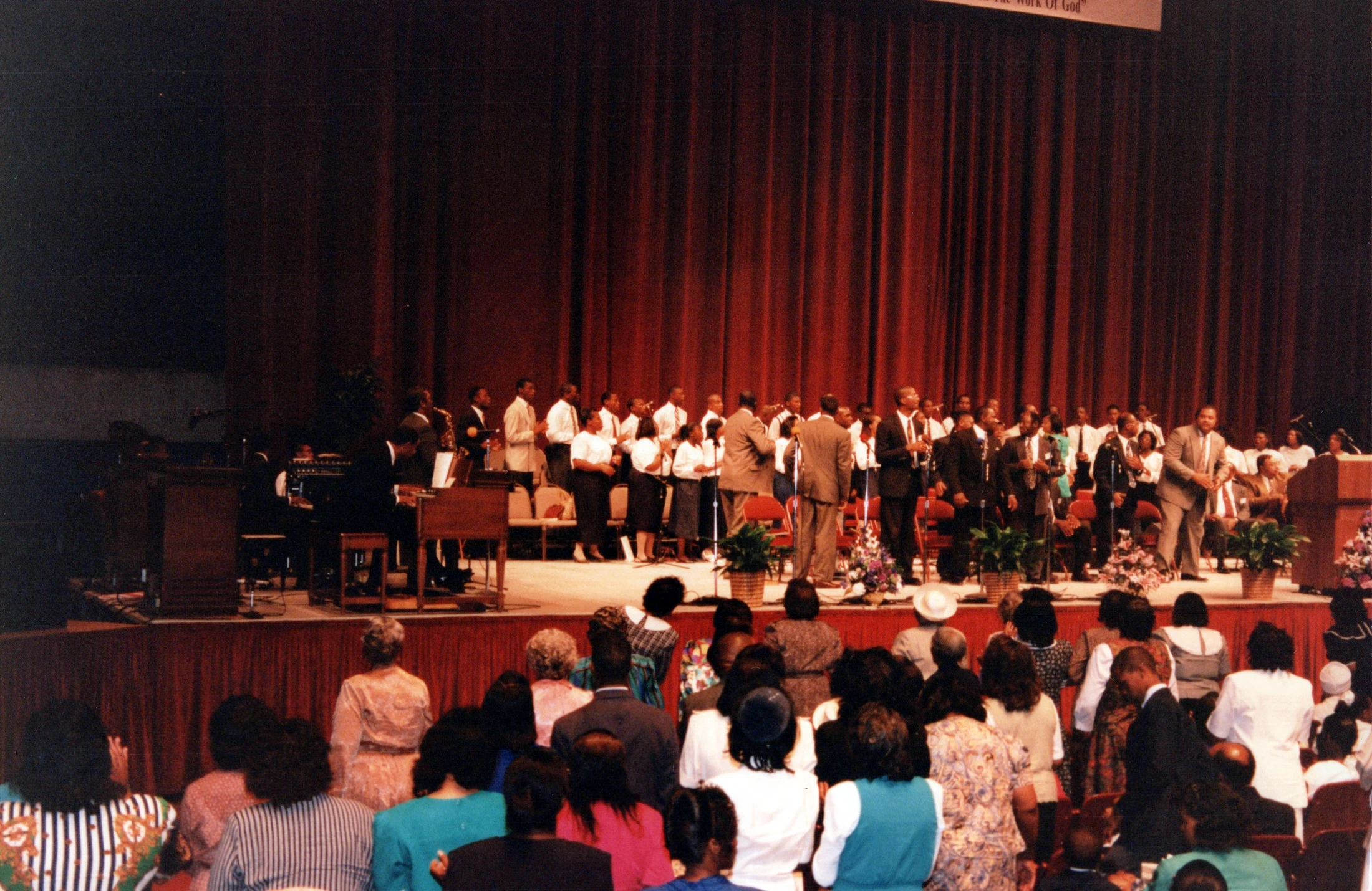 An African American choir and musicians perform on stage in front of a red curtain for a large audience in an auditorium.